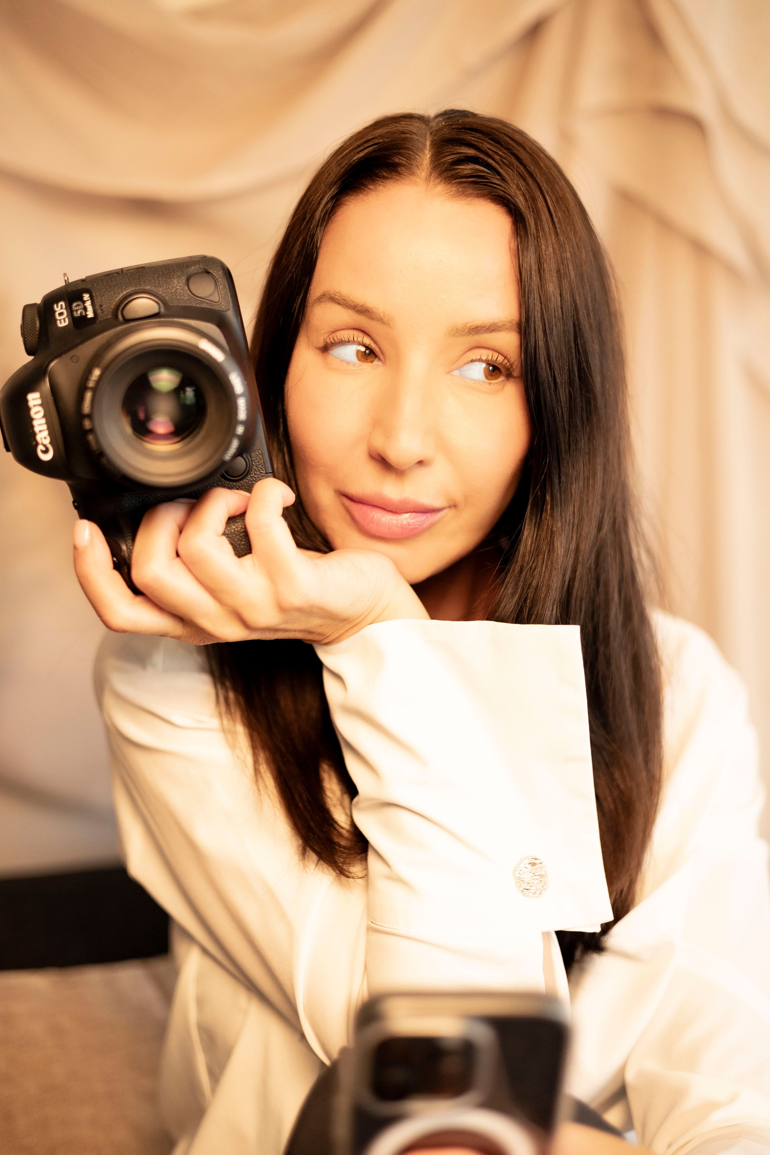 bild på fotografen Sabina som håller en systemkamera i handen. i bakgrunden syns vit tyg och i förgrunden syns en mobil.