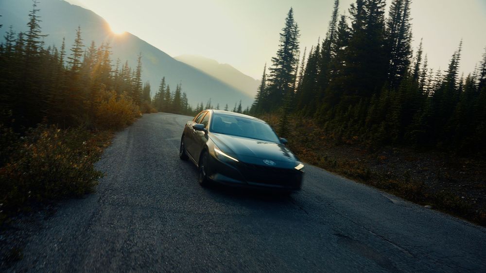 Dark sports car driving on a mountain road surrounded by evergreen trees at dusk.