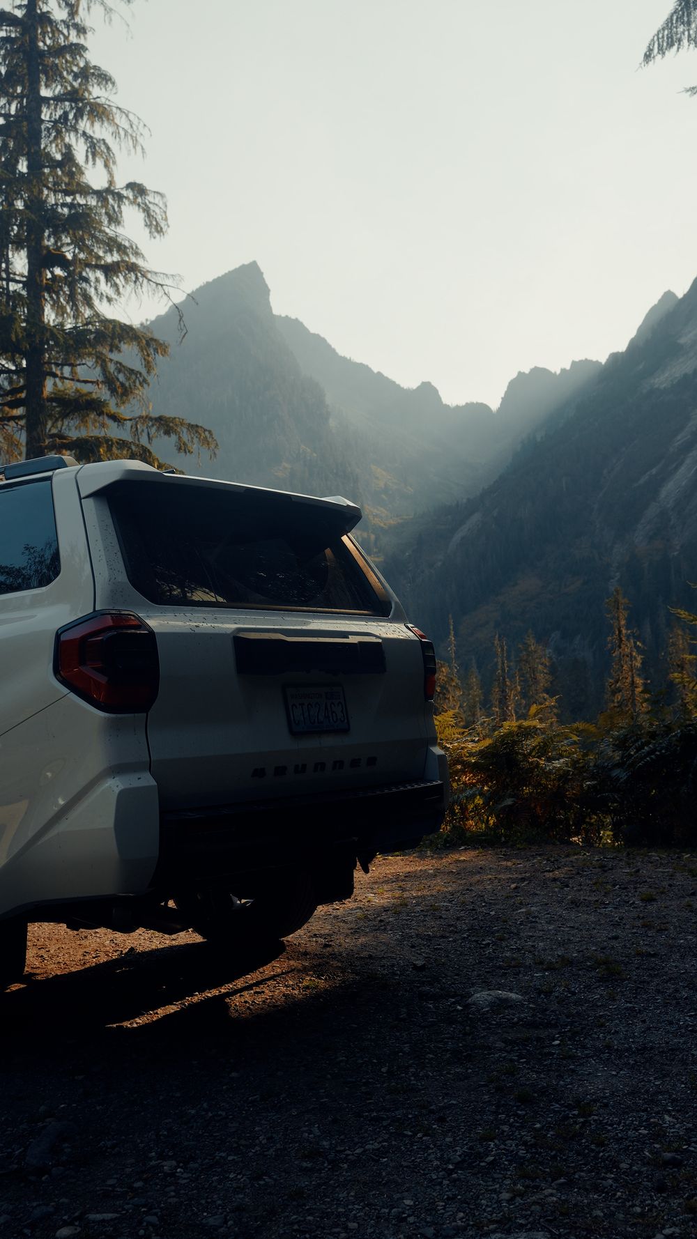 White SUV parked on mountain trail with dramatic mountain peaks in background during golden hour.
