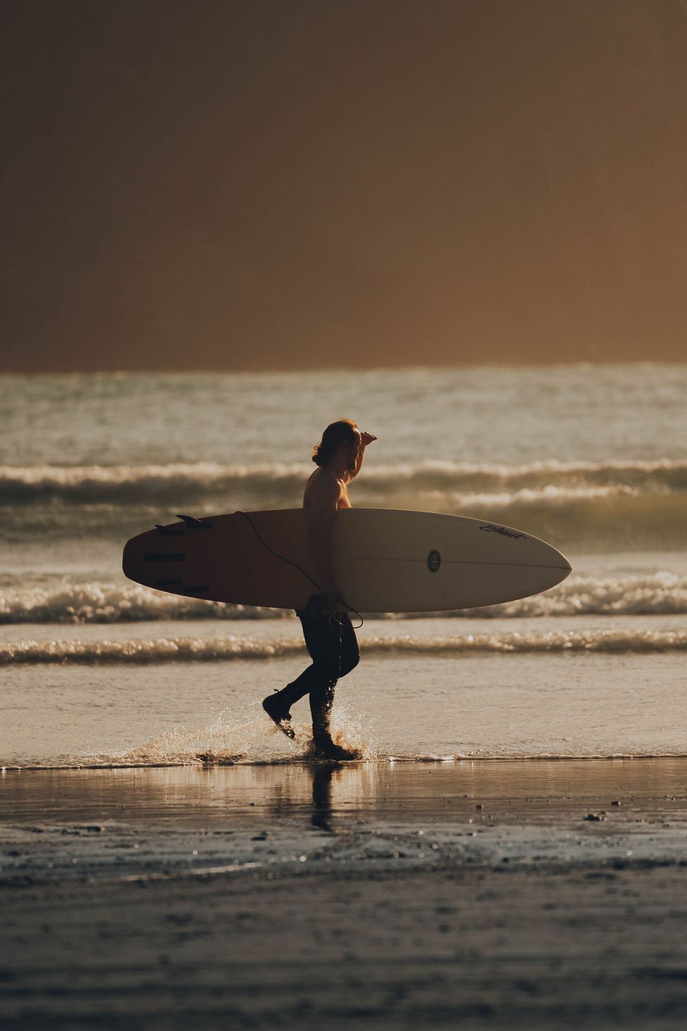Silhouette of surfer walking on beach at sunset carrying surfboard.