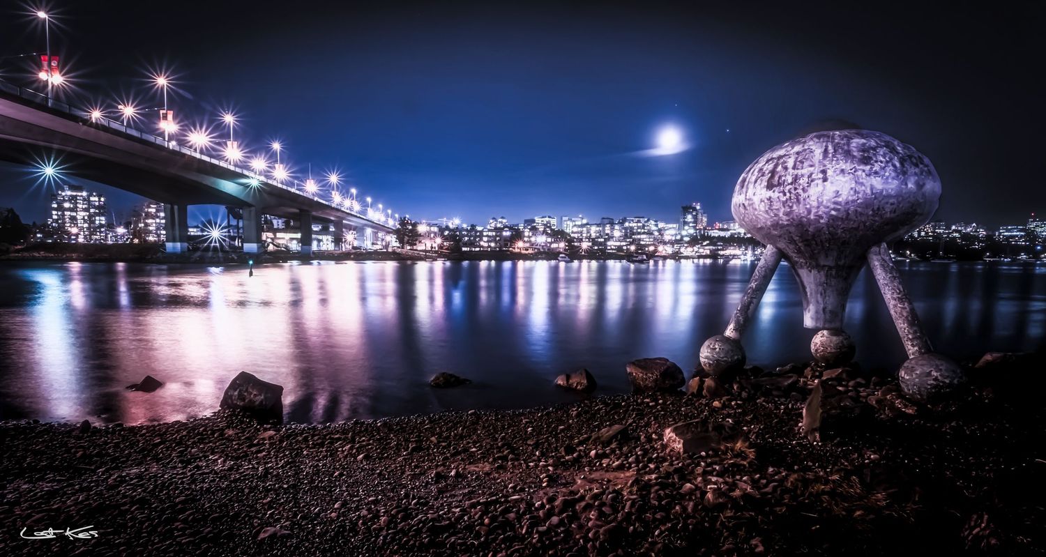 Glowing mushroom sculpture illuminated at night along Vancouver's waterfront with Cambie Bridge and city lights reflecting.