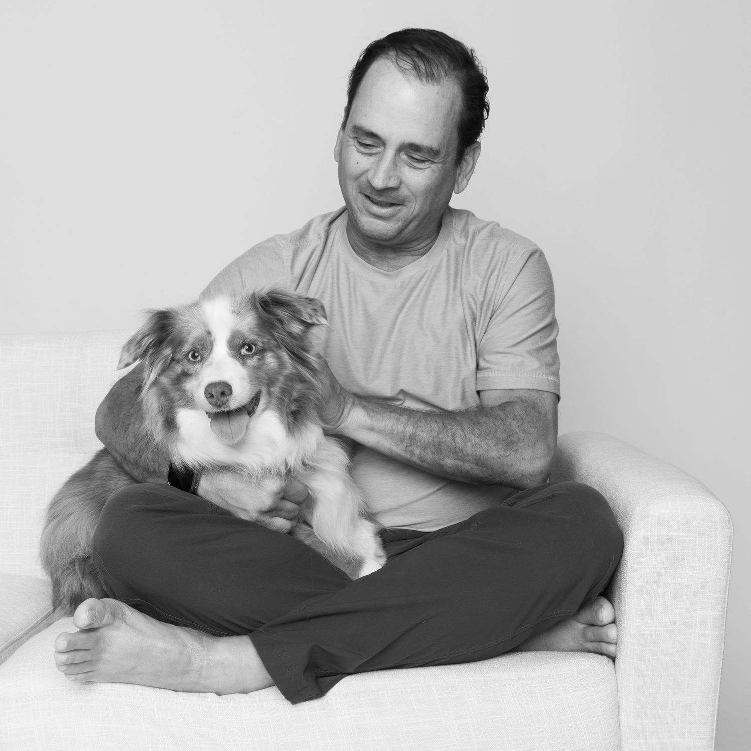 A person sits cross-legged in casual attire with a smiling Australian Shepherd dog in a black and white portrait.