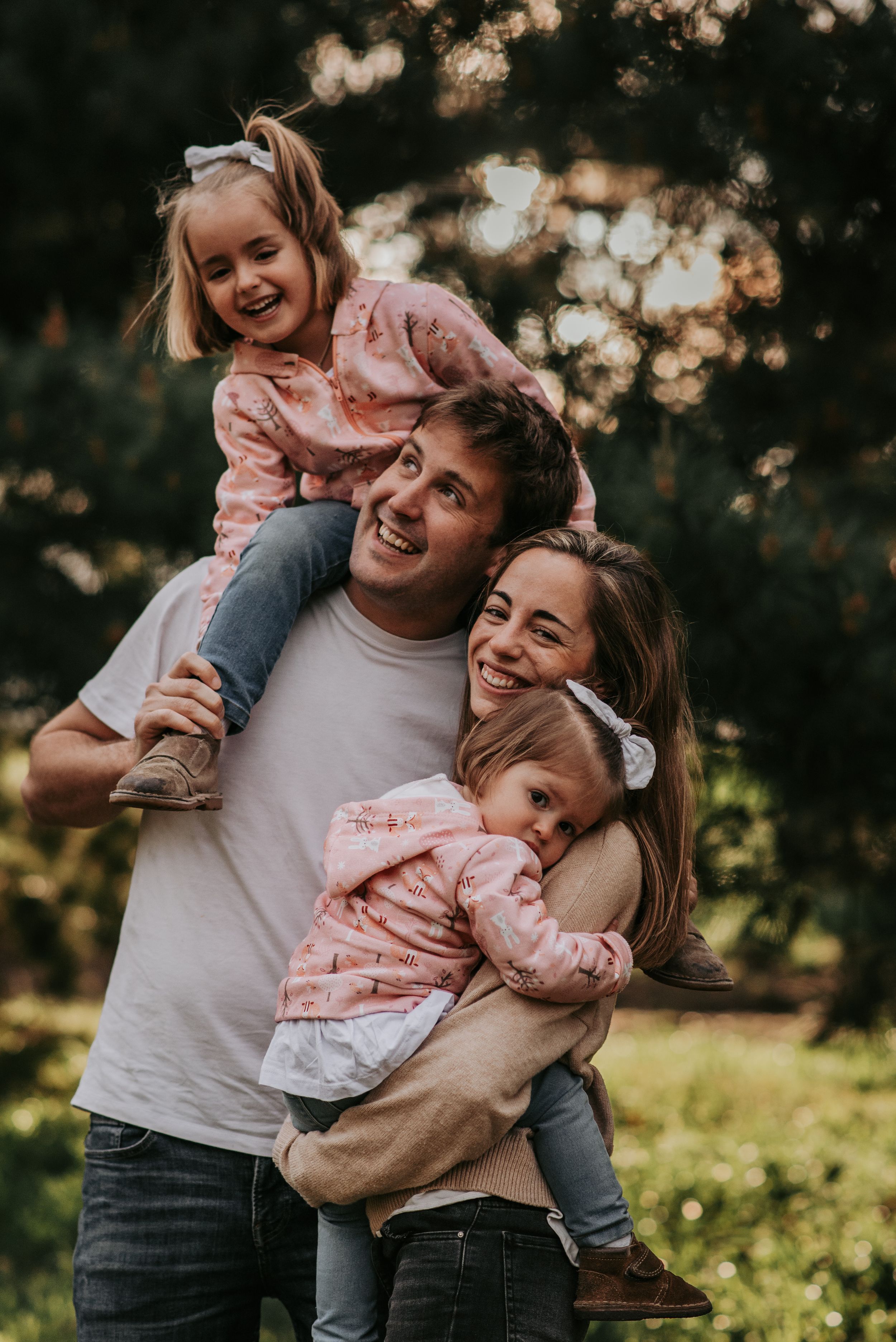 Foto familiar, niña de 5 años sobre los hombros de su padre sonriendo, niña de dos años a upa de su madre mira tímida