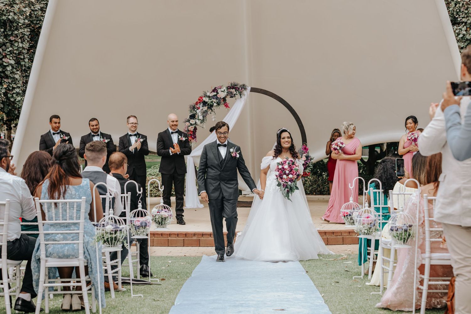 Bride and groom walking down the aisle at outdoor wedding ceremony with floral arch.