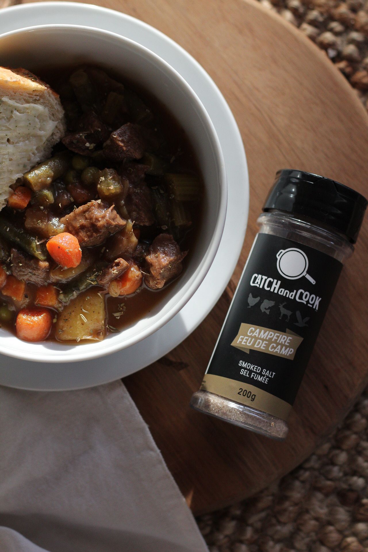 Bowl of hearty stew with vegetables next to a Catch and Cook seasoning spice jar on wooden surface.