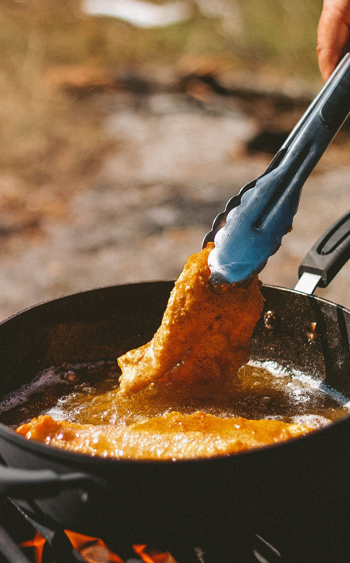 Blue tongs stirring bright orange sauce in a cast iron pan over outdoor flame.