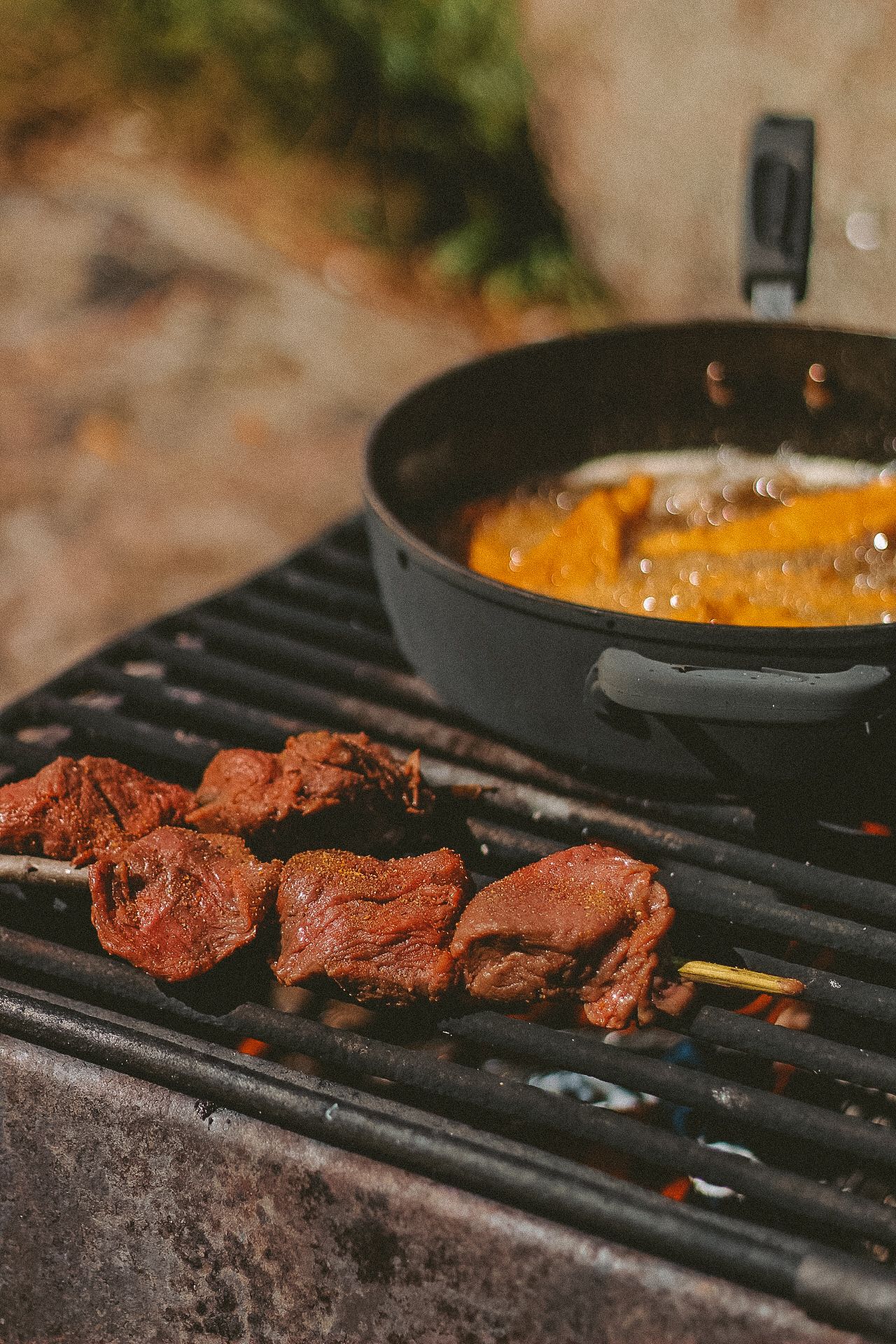 Marinated meat skewers grilling on barbecue next to pan of orange sauce.