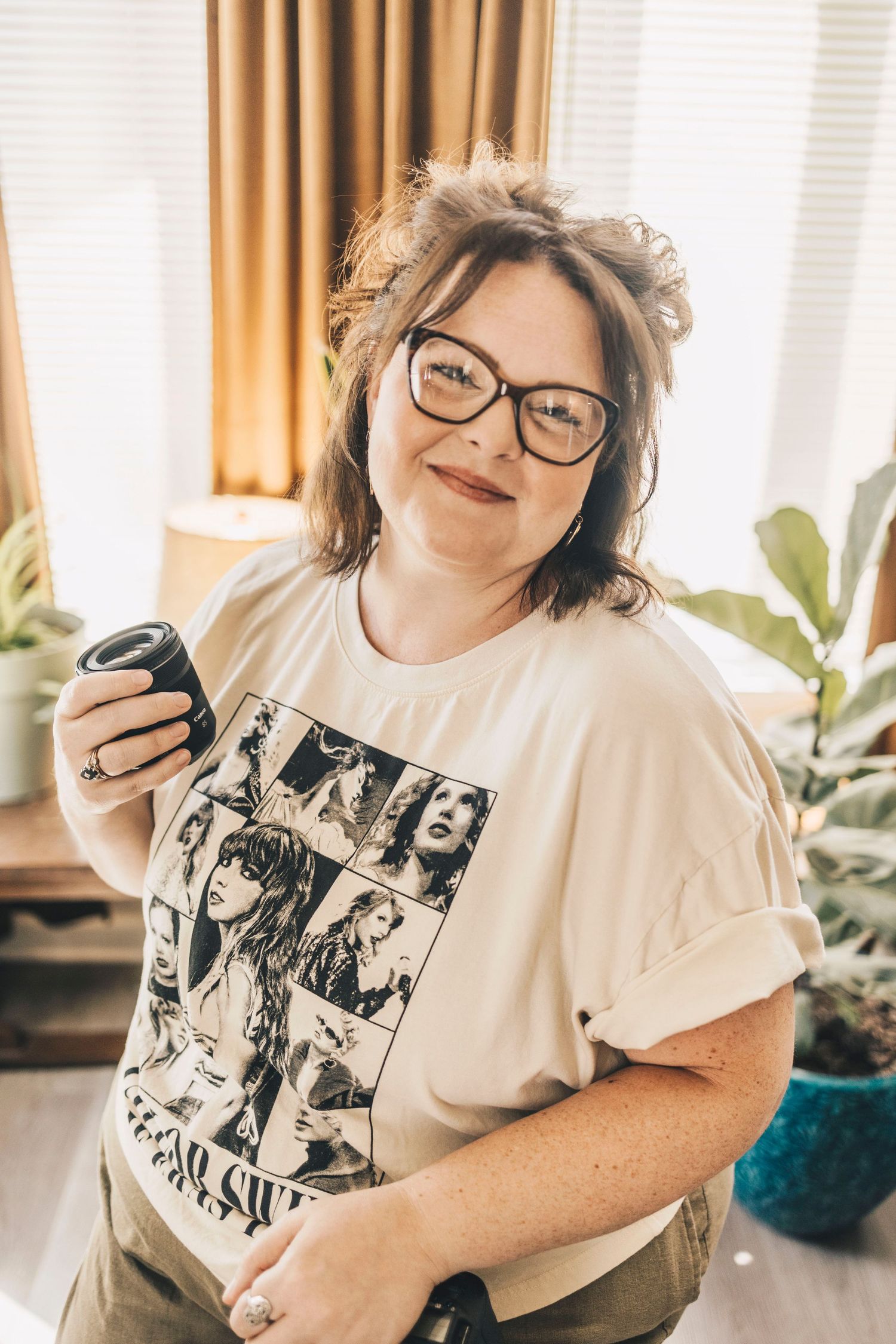 Woman in Taylor Swift Eras Tour shirt holding camera lens smiling near window with plants.