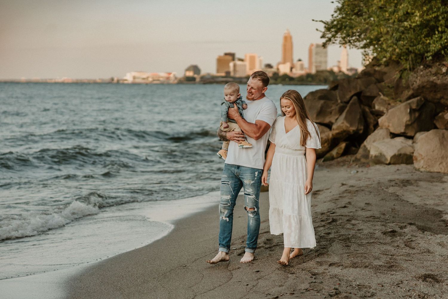 Family walking along rocky beach shore with city skyline visible in background.