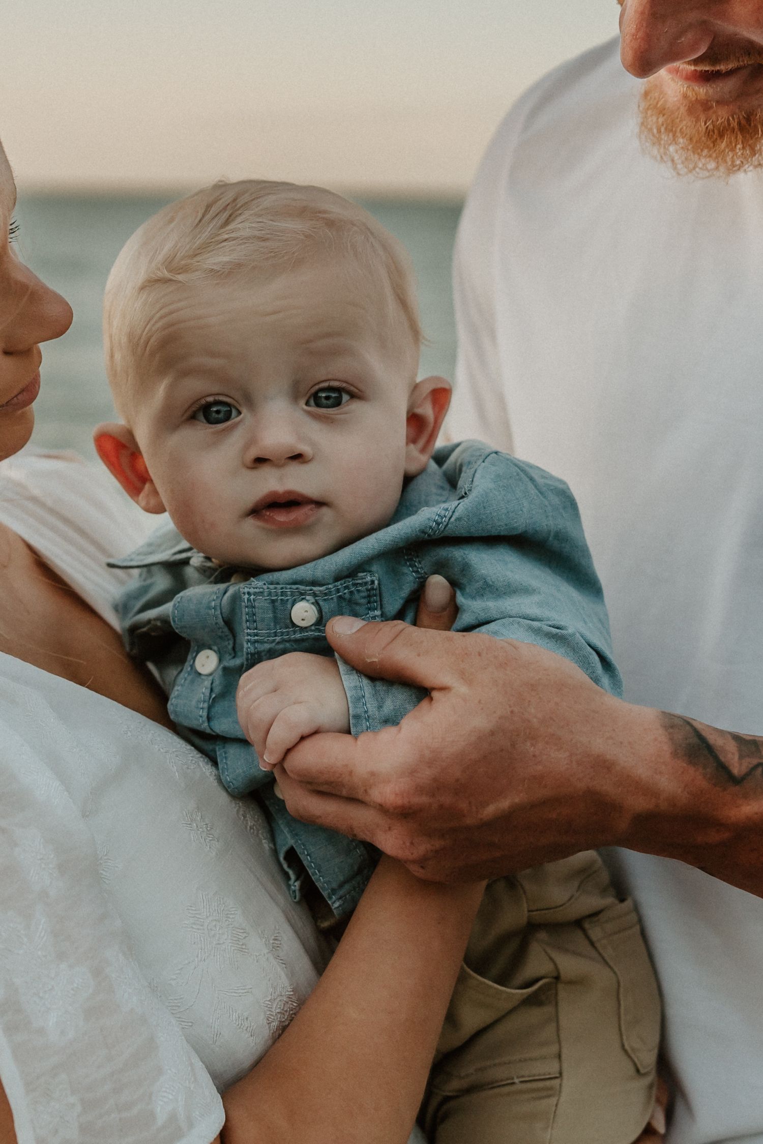 A small baby in denim clothing is held lovingly by two adults against a soft sunset background.