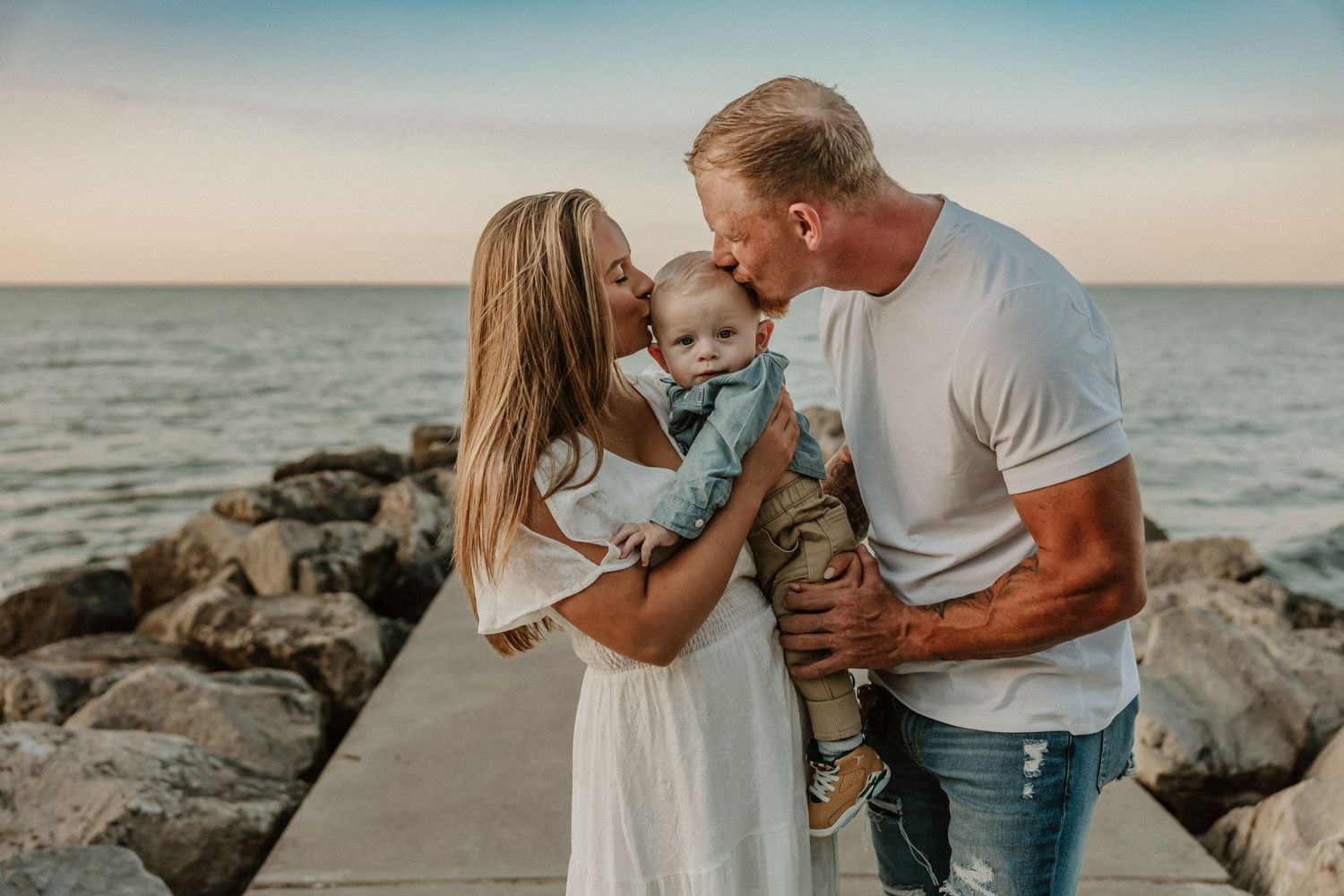 A family shares an intimate moment together on a rocky pier by the ocean at sunset.