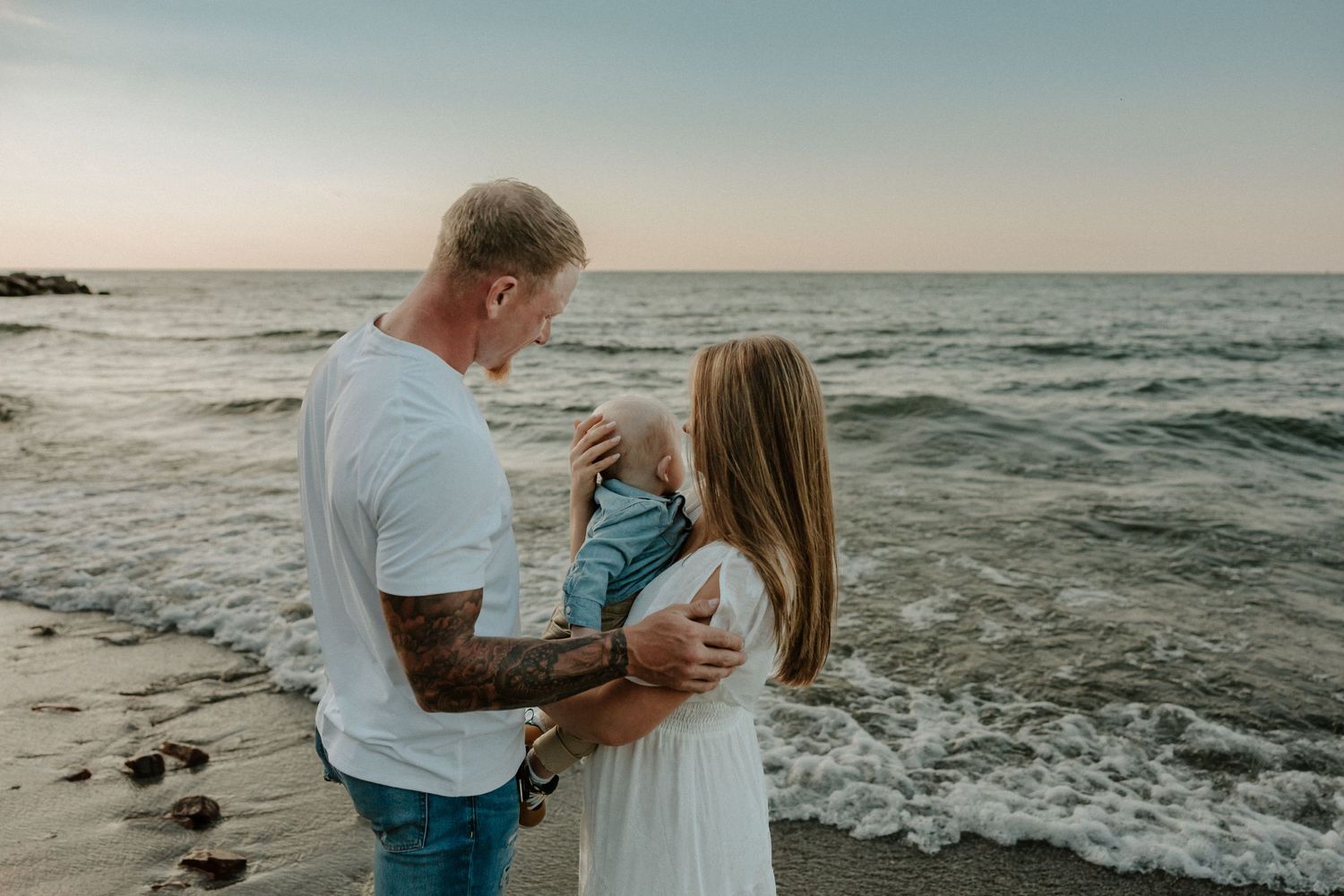 A family embraces while standing on a coastal pier with waves crashing against rocks at sunset.