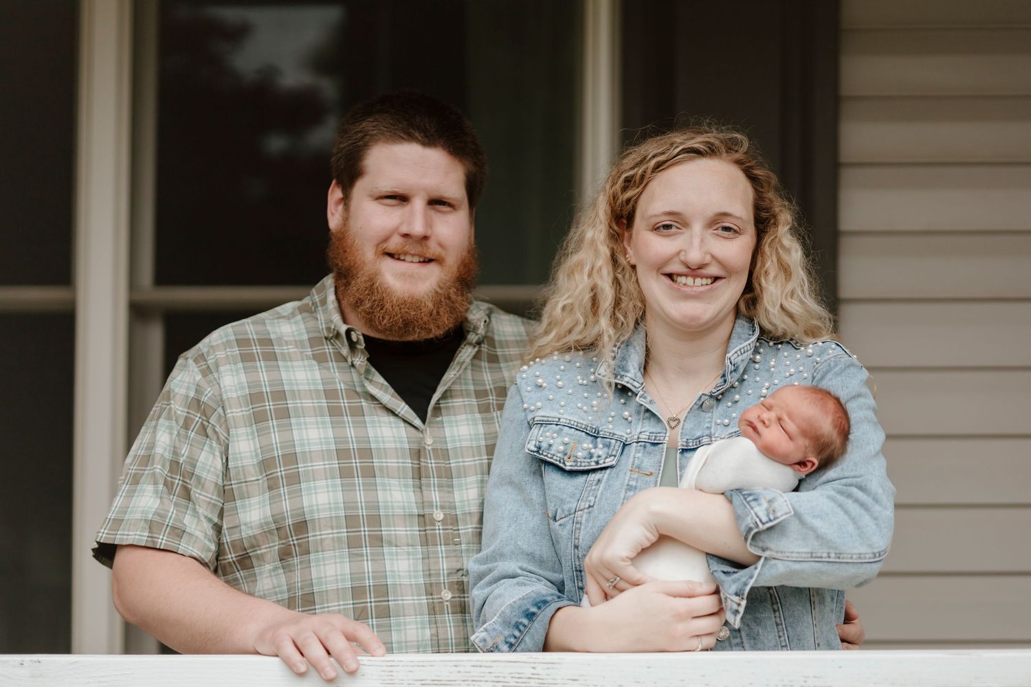 A family poses together on their front porch while holding their newborn baby.