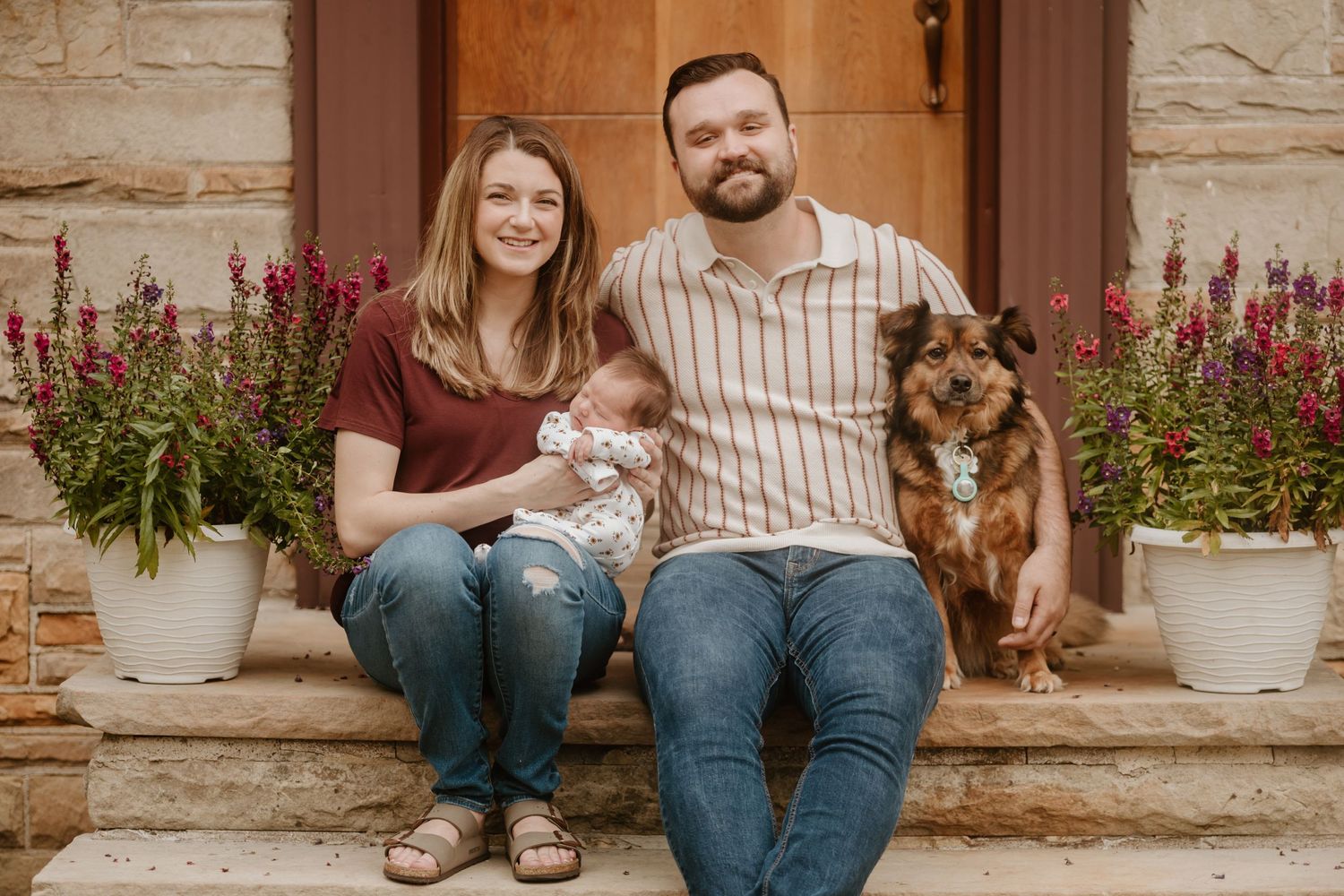 A family sits on outdoor steps with potted flowers and their dog while holding a small baby.