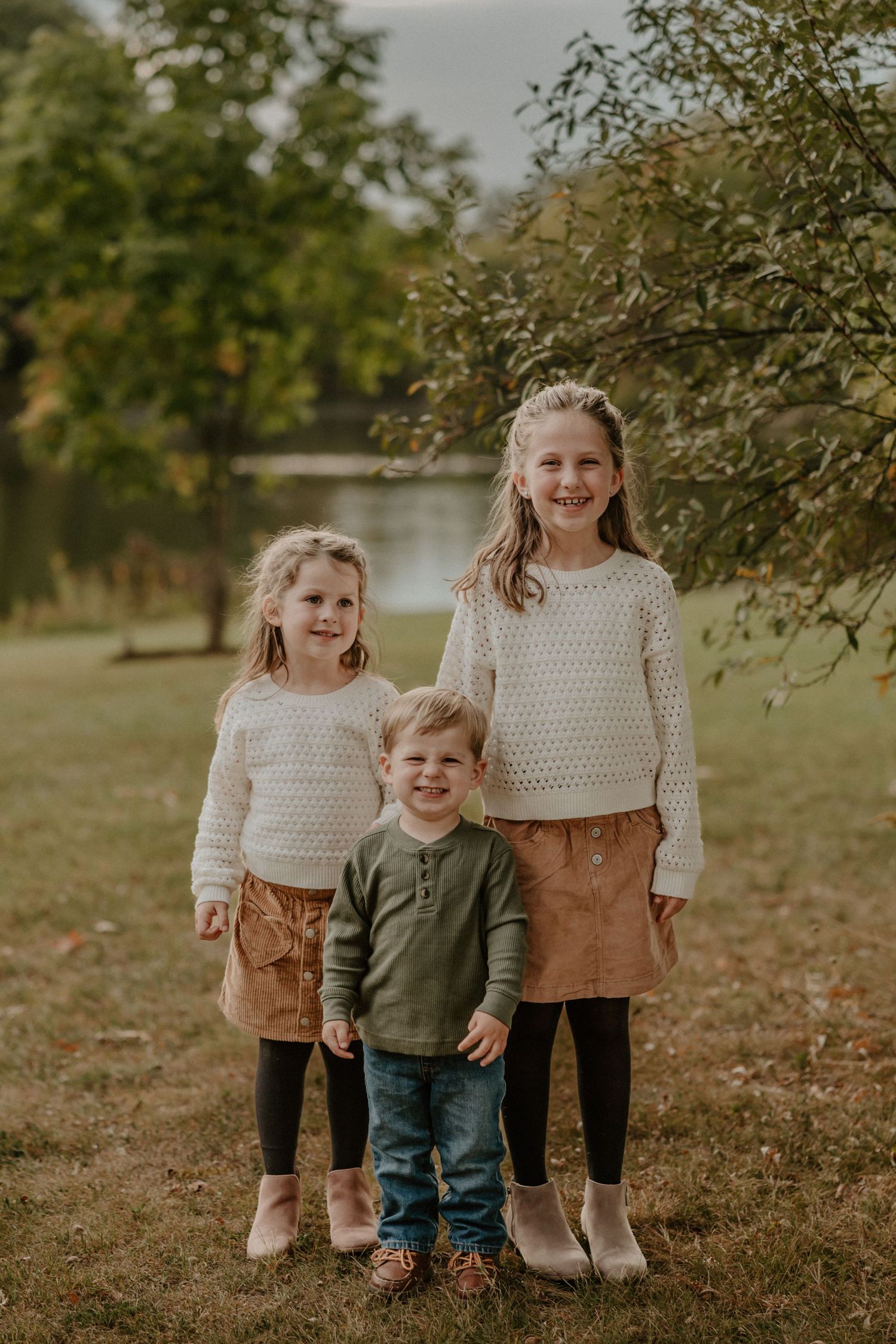 Three siblings in cozy sweaters pose together for a fall portrait by a lake.