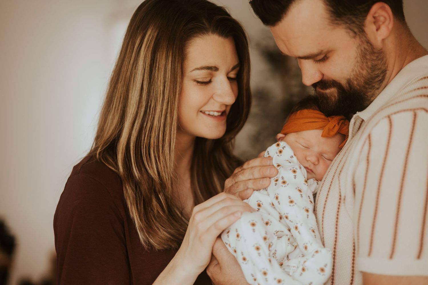 A couple gazes lovingly at their newborn baby wrapped in a floral print blanket in a warm indoor setting.