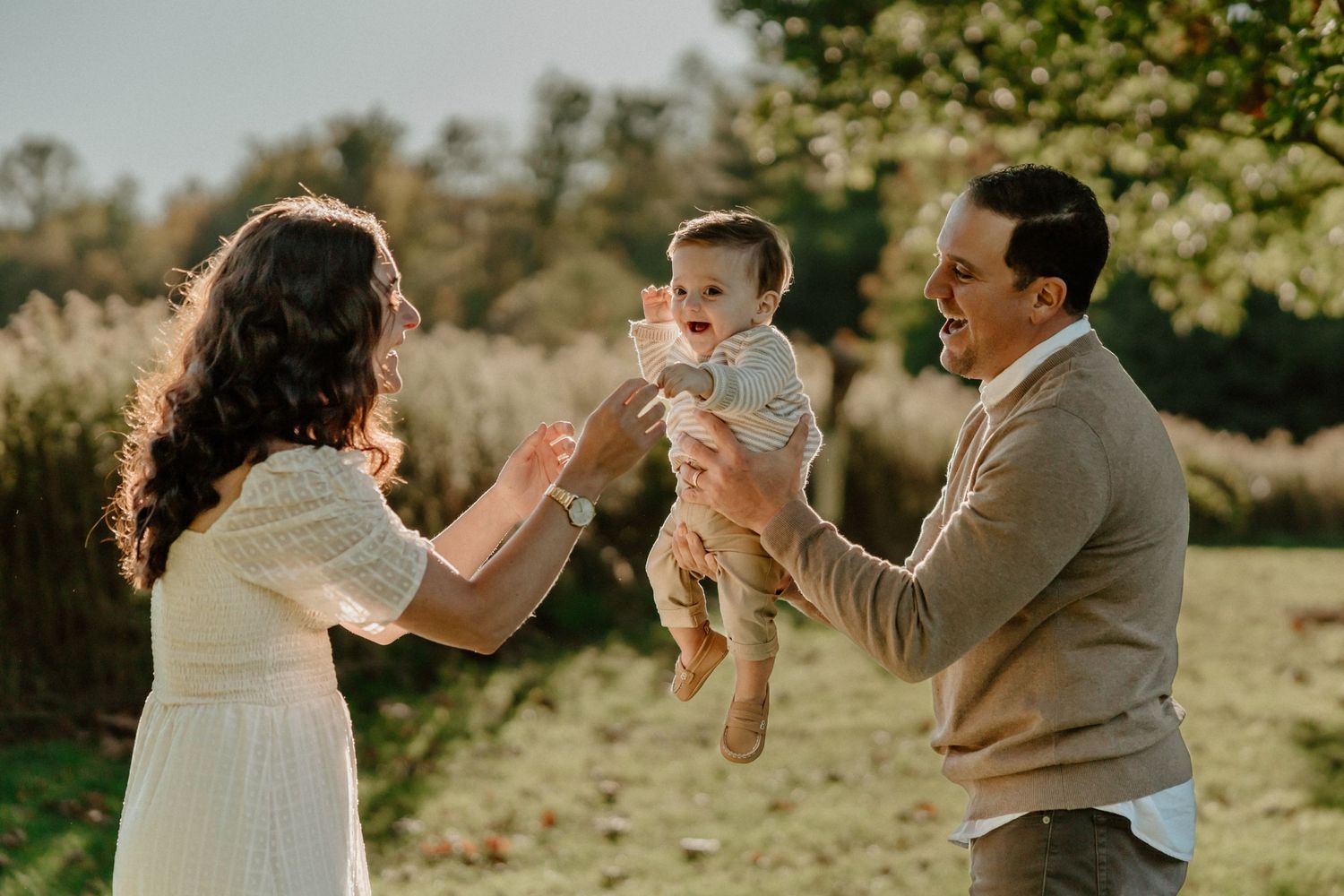A family plays together in a sunlit field, lifting their baby in a joyful moment.