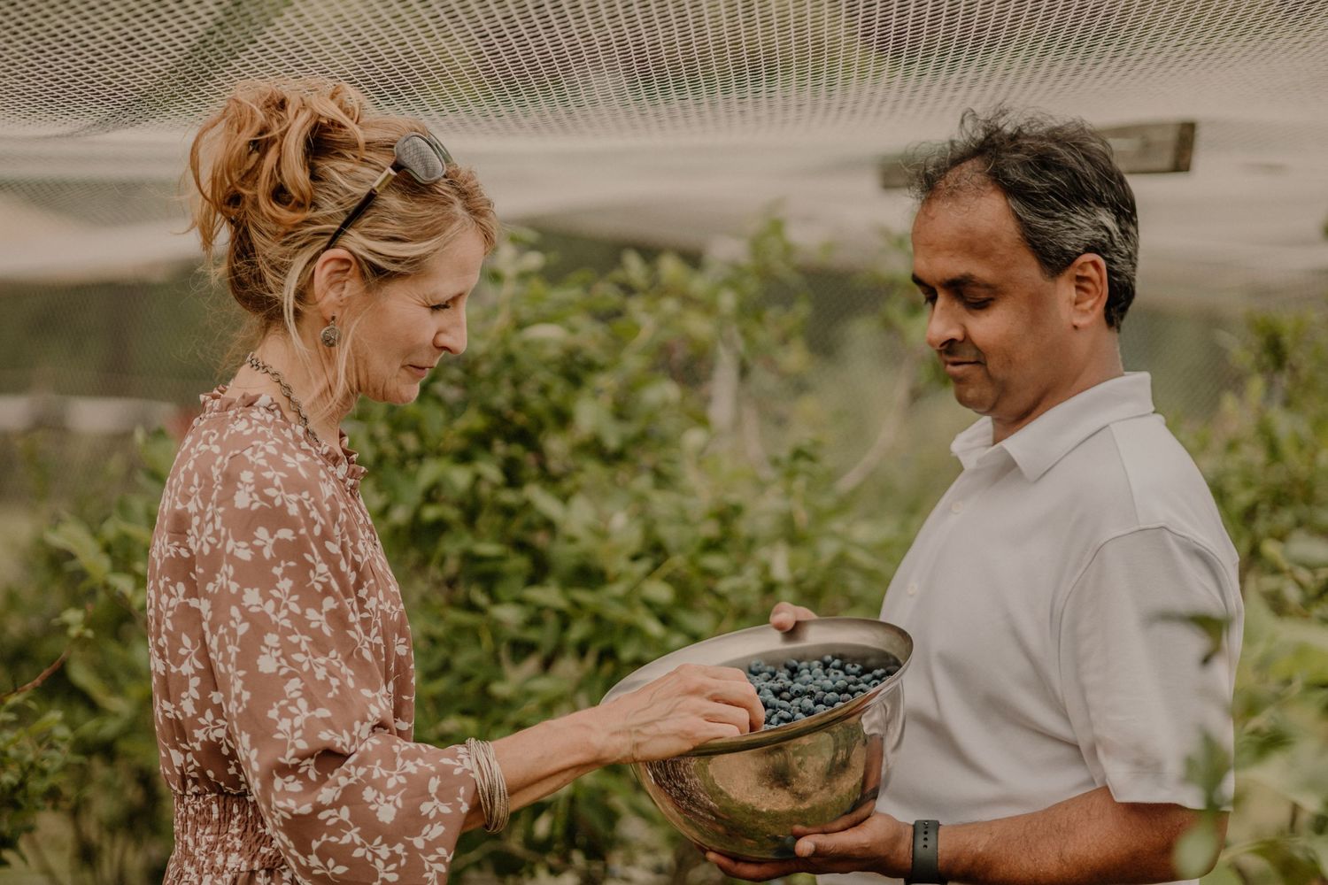 A couple examines fresh berries in a bowl while standing in a garden.