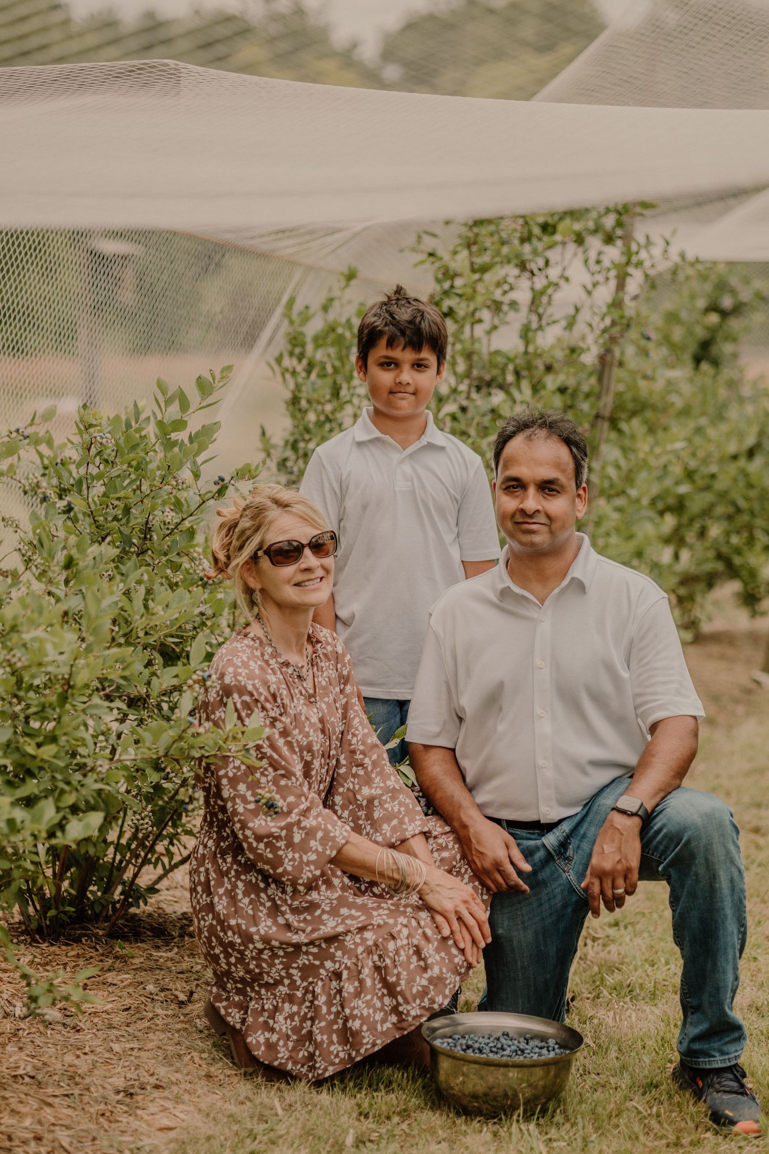 Family gathered together amongst blueberry bushes on their farm under protective netting on a sunny day.