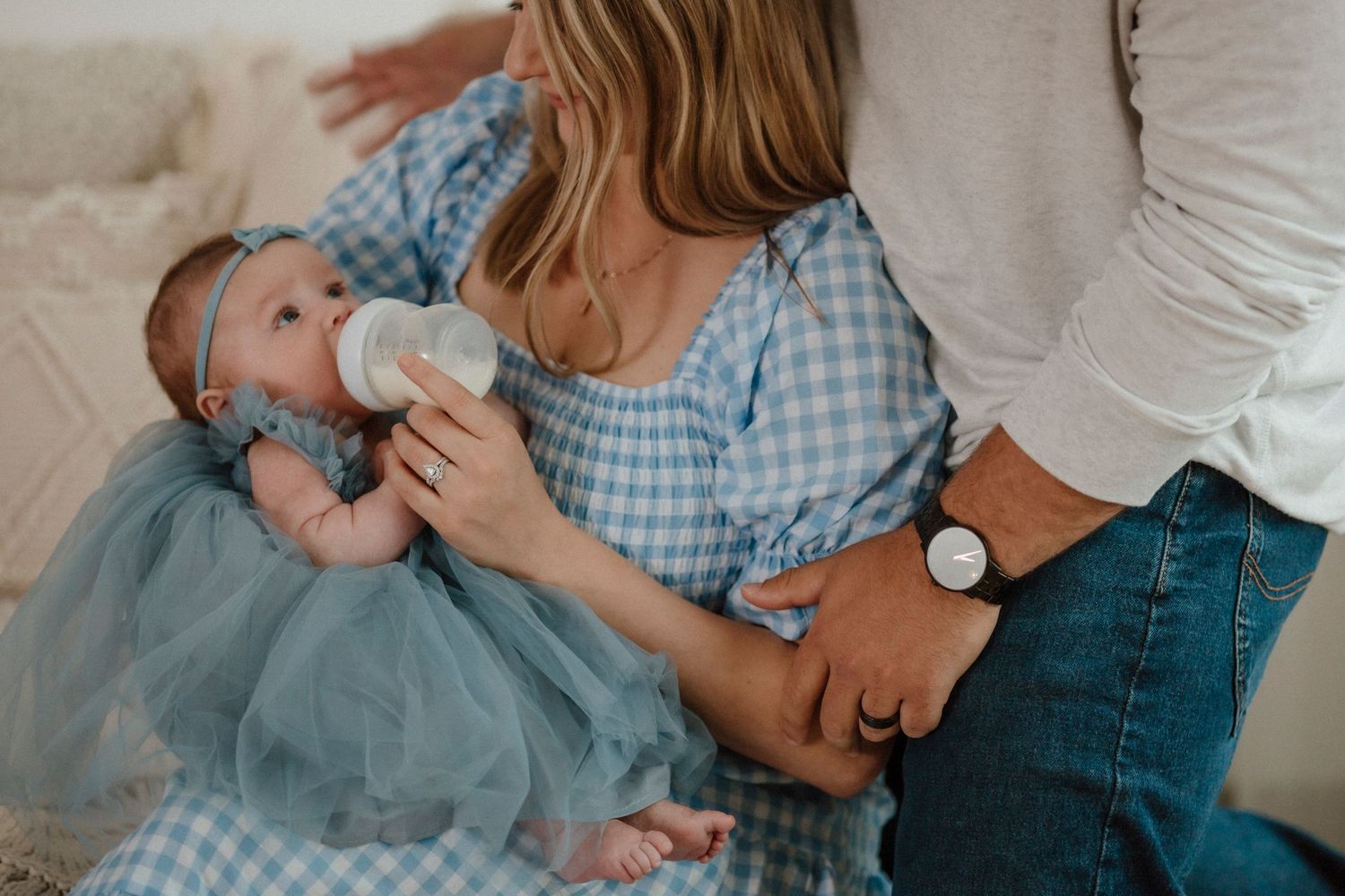 Baby in blue tulle dress being bottle fed while parents share an intimate moment.