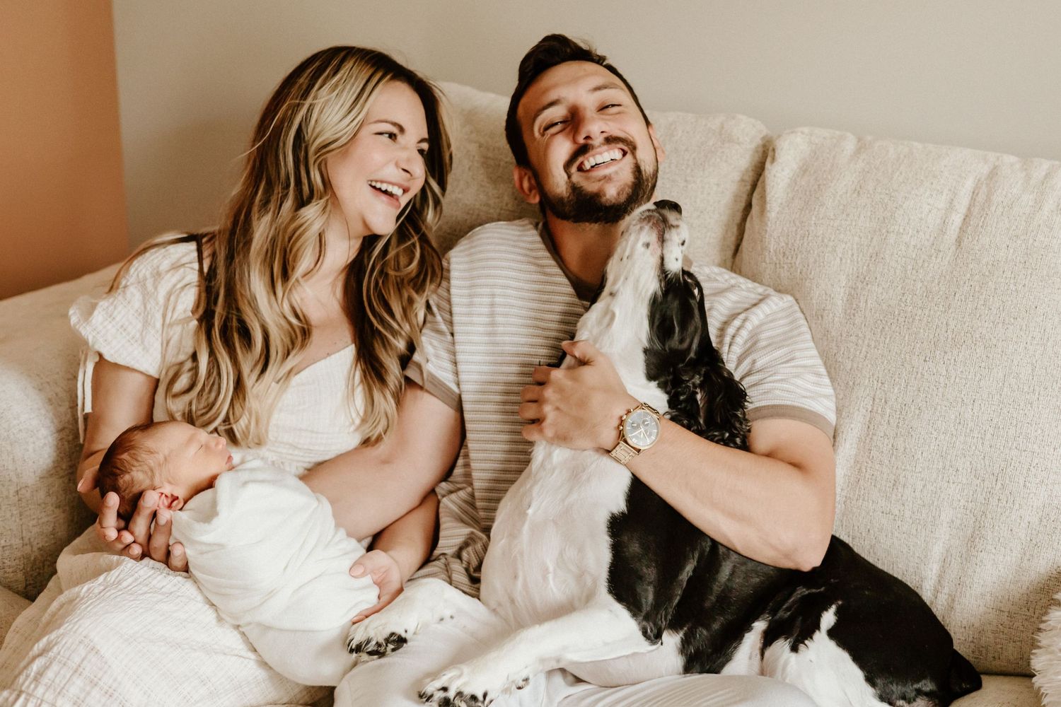 Family shares joyful moment on couch with their black and white dog and baby.