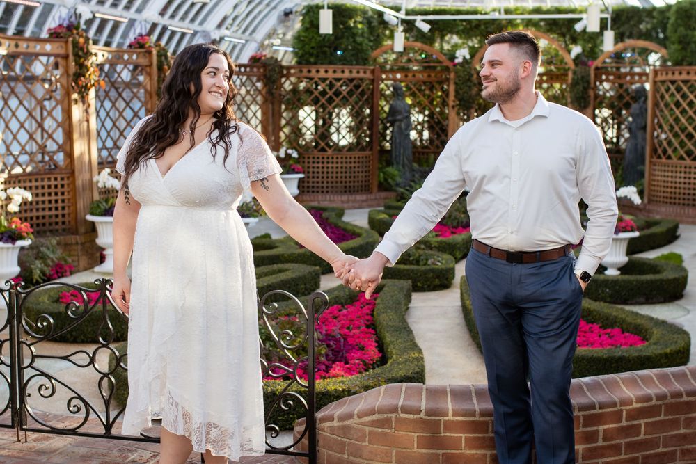A couple holds hands in a garden setting with blooming flowers and lattice decorations, wearing white dressy attire.