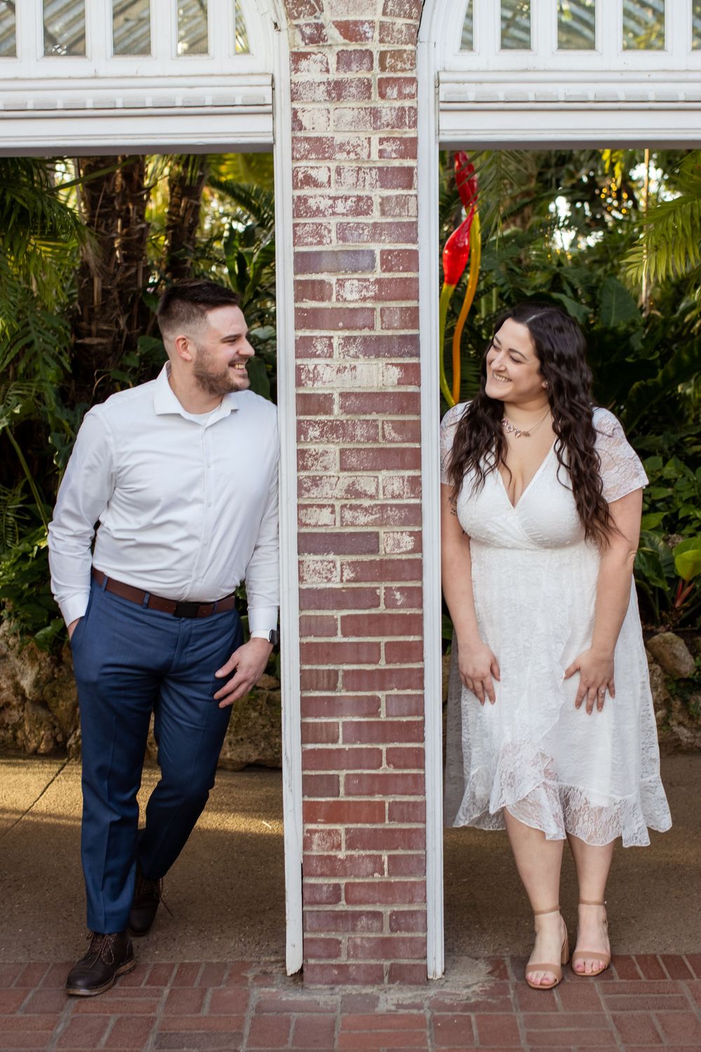 A couple poses playfully on either side of a brick column in a garden patio setting, wearing coordinating white outfits.