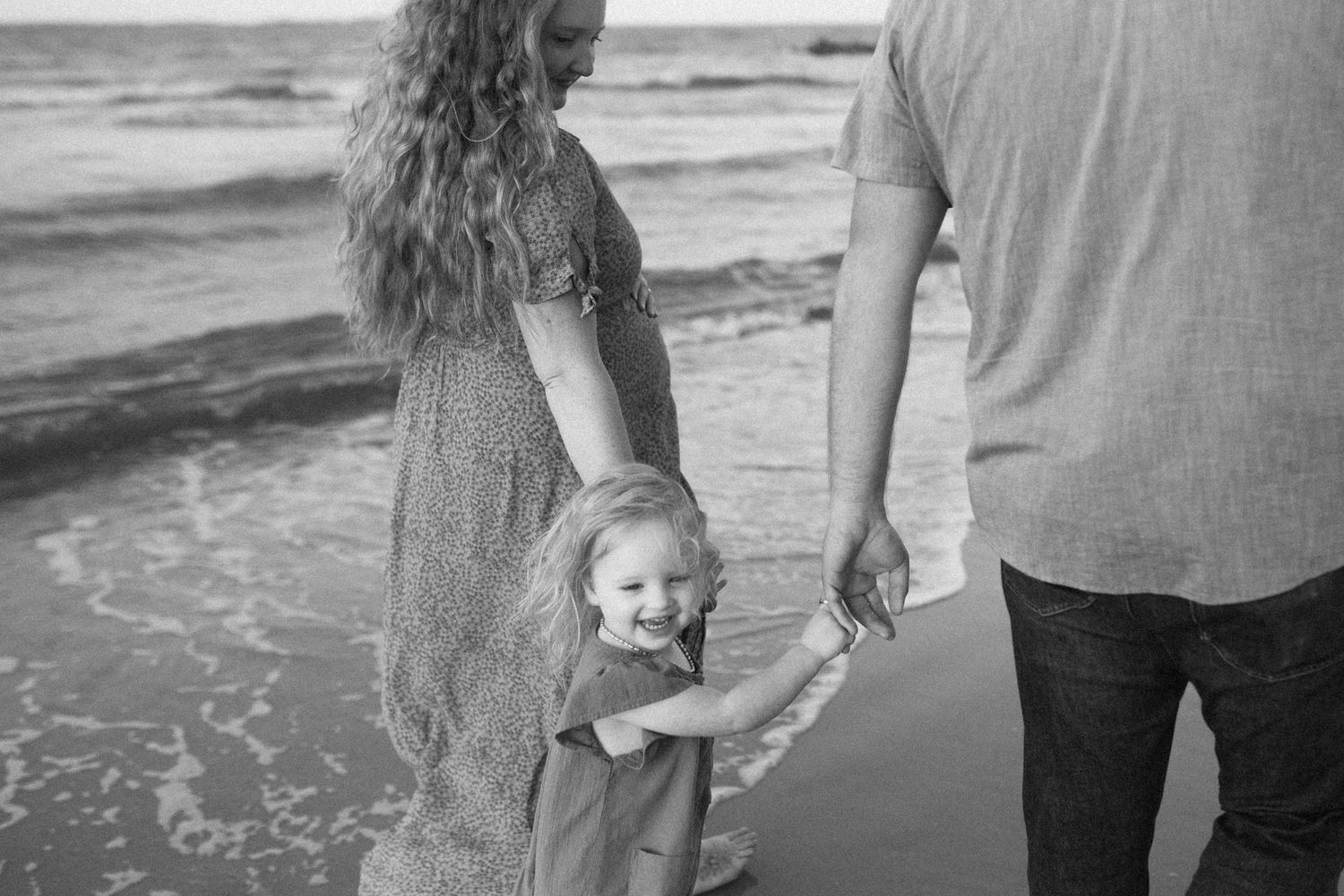 A family holds hands while standing at the edge of the ocean waves on Hilton Head Island captured in black and white by B. Logsdon.