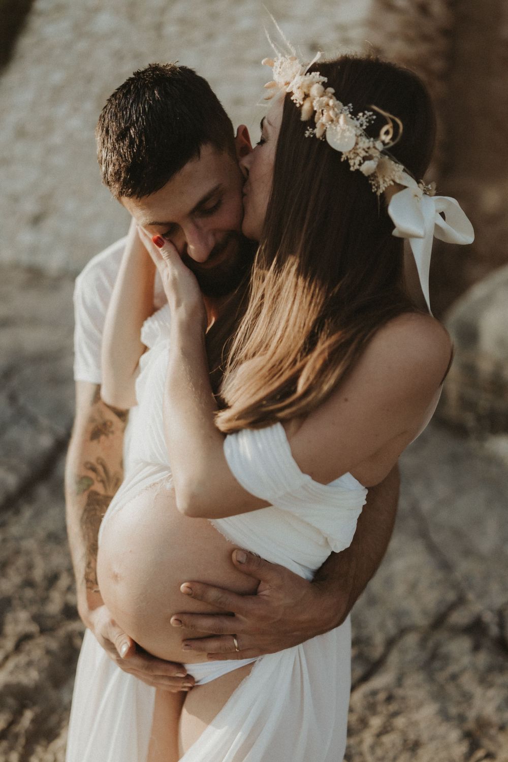 Intimate maternity portrait with flower crown and white dress against rocky coastal backdrop.