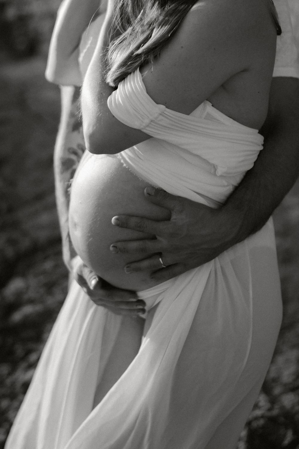 Black and white intimate maternity photo showing gentle embrace and flowing white dress.