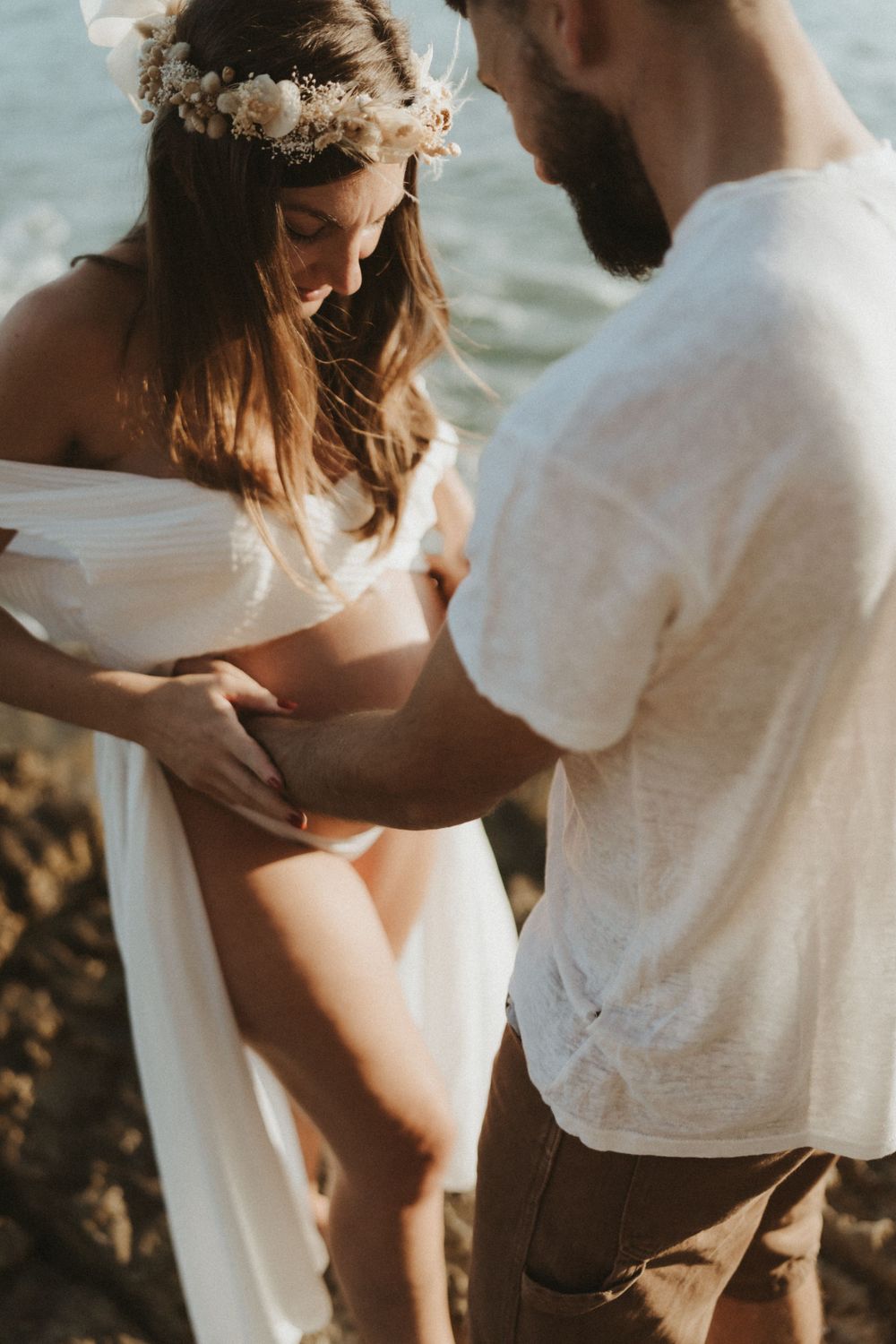 Sun-drenched maternity photo capturing tender moment between couple on coastal rocks.