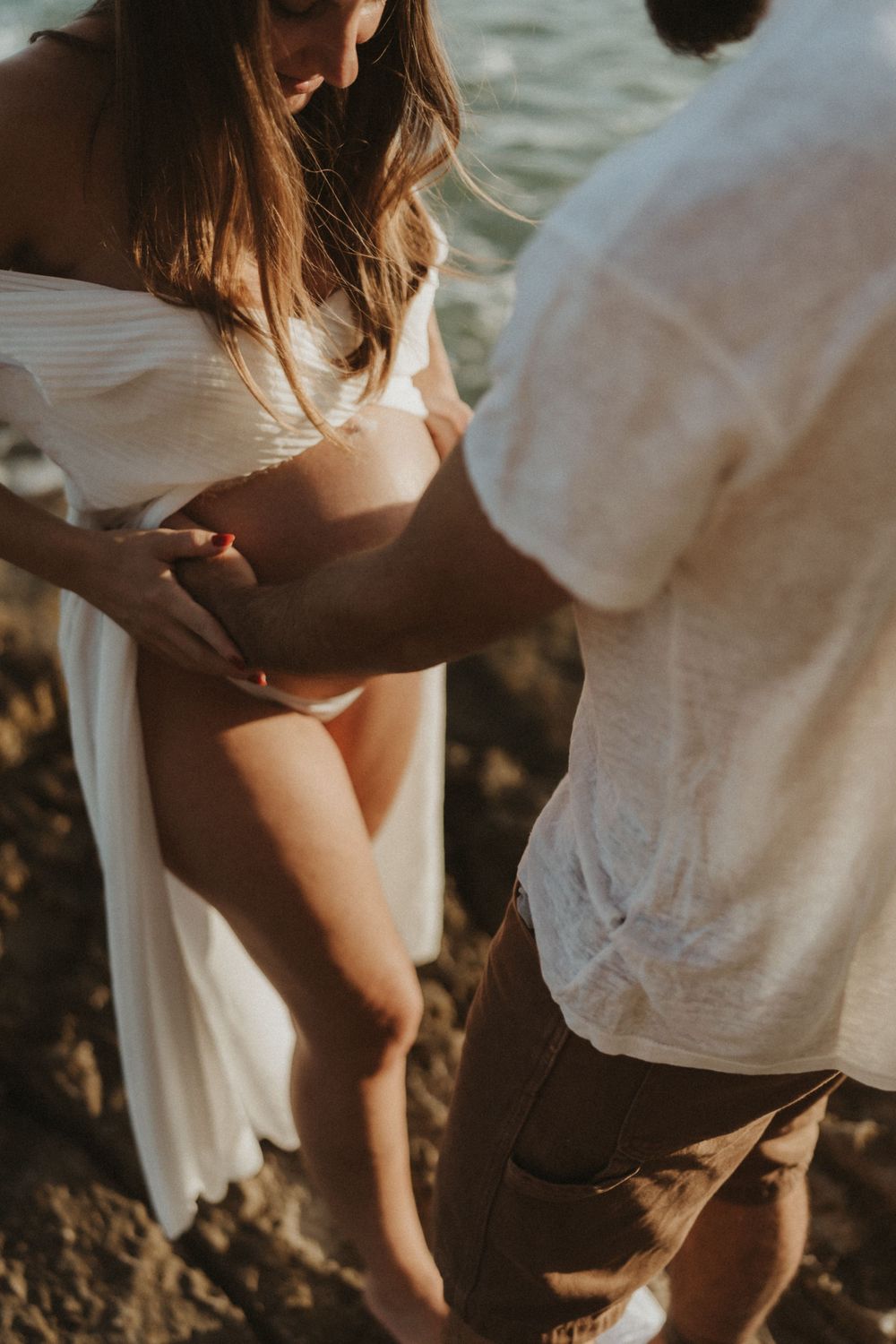 Intimate sunset maternity photo of couple holding hands on rocky beach in soft golden light.