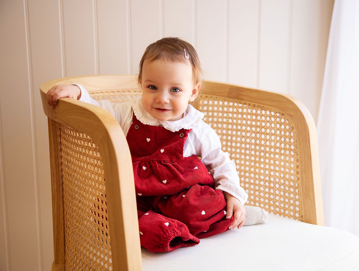 A small child wearing a festive red jumpsuit sits happily in a light wooden cane back chair. During a baby photoshoot in Bexley