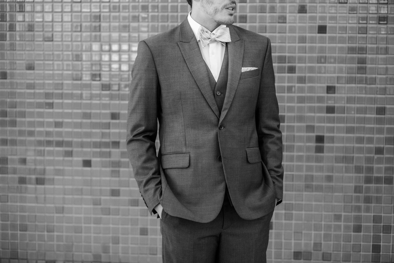 Classic black and white portrait of groom in formal suit against tiled wall.