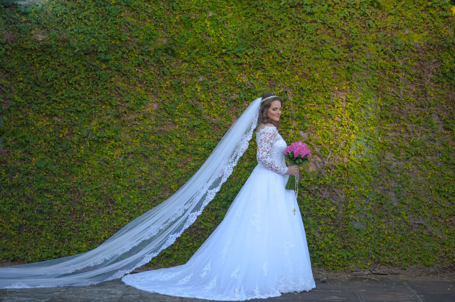Aerial view of bride in flowing white dress and veil against lush green garden wall.