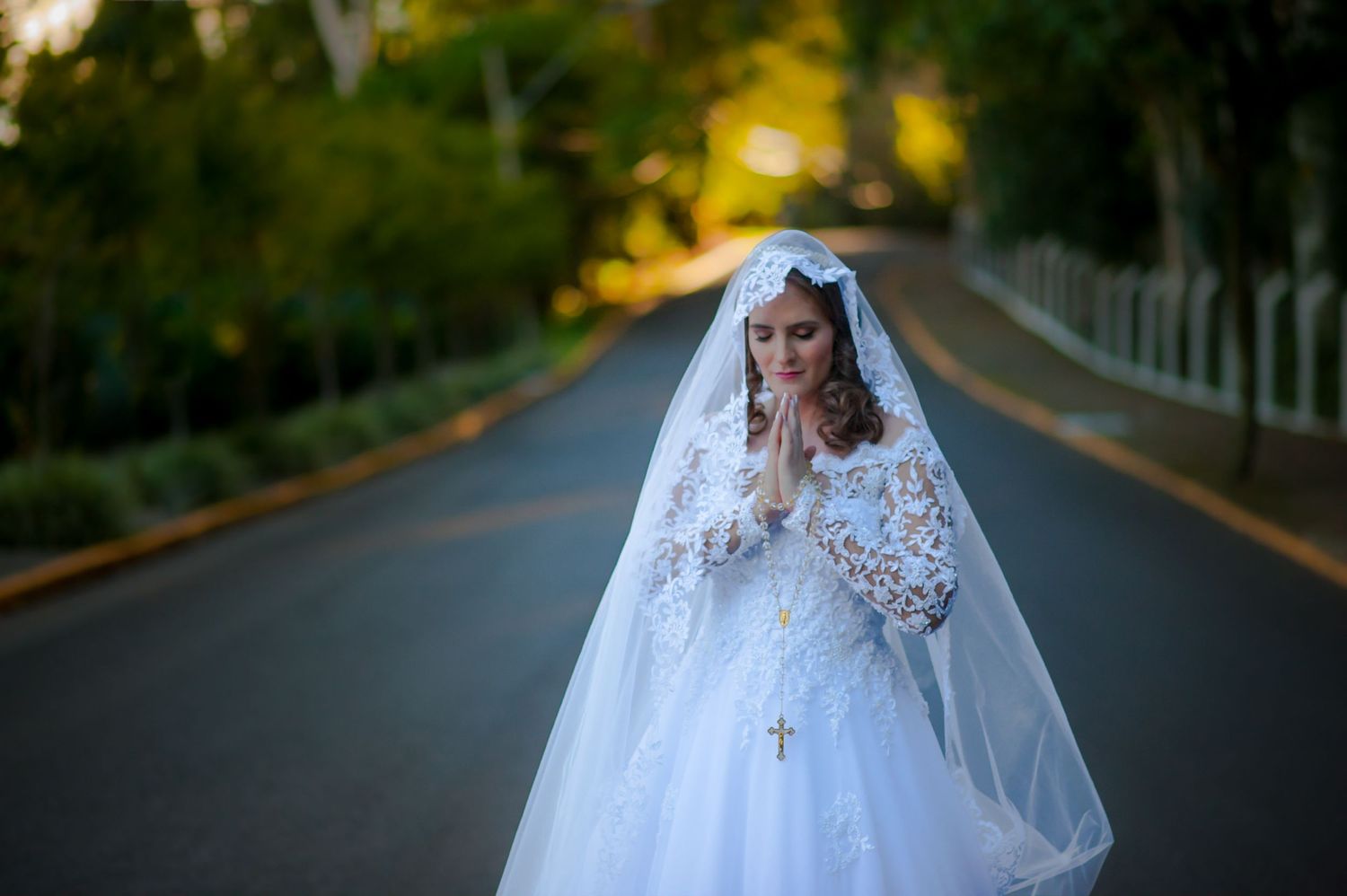 Bride in white lace gown and veil standing on tree-lined road at dusk.