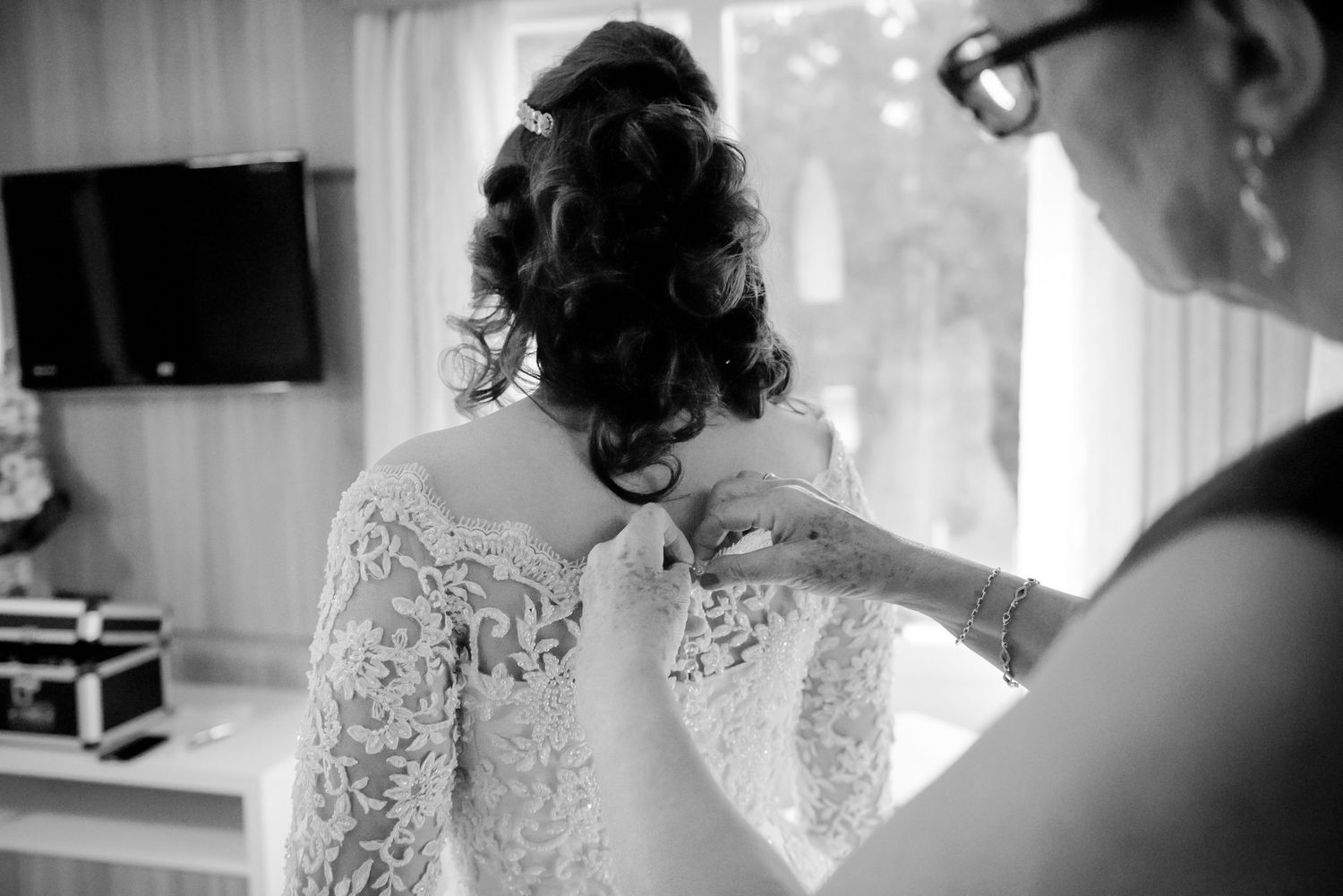 A delicate lace wedding dress being fastened in a black and white preparation scene.