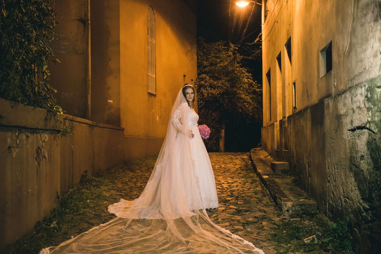 Bride in flowing pink gown stands in atmospheric alleyway at night.