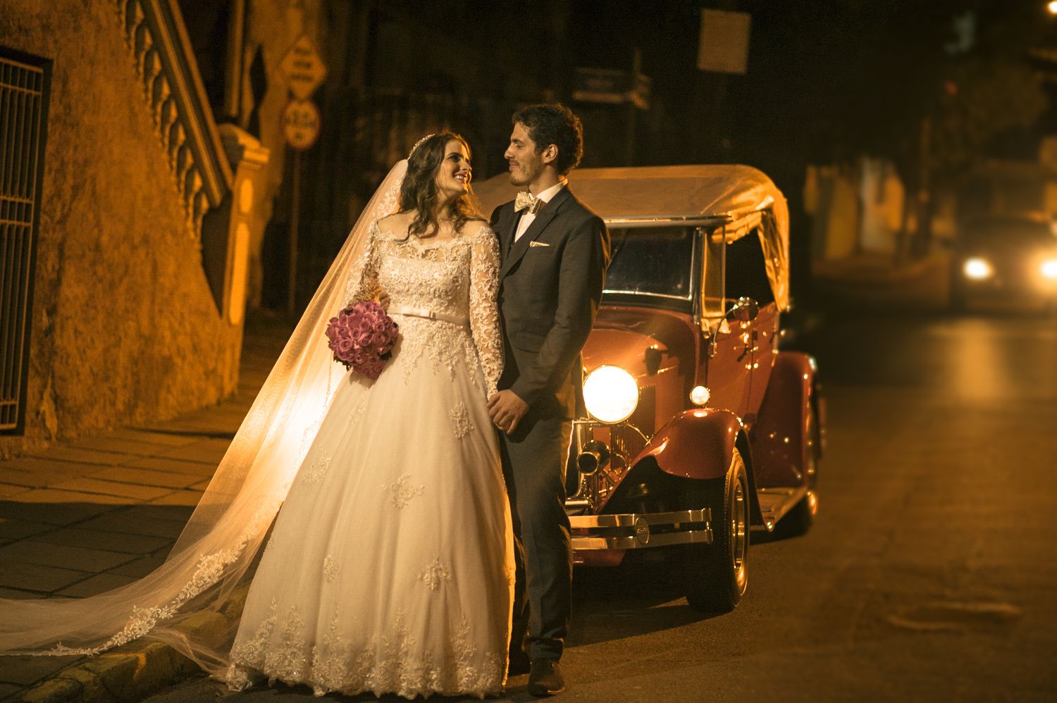 Wedding couple poses near vintage car on cobblestone street at night.