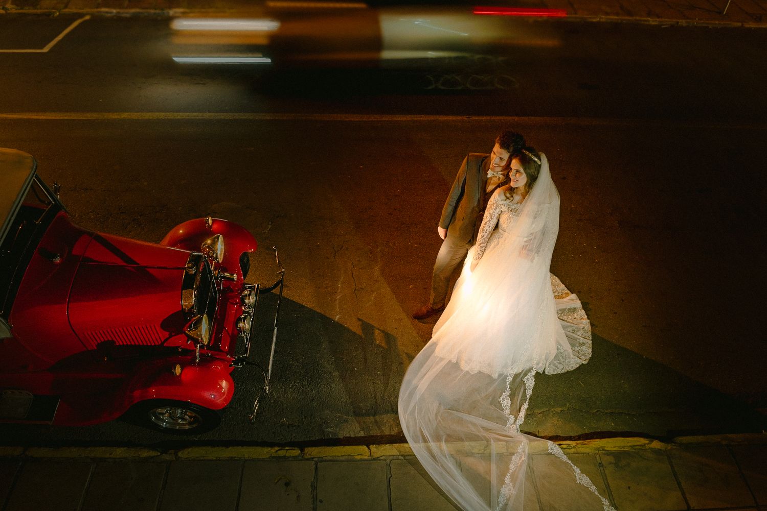 Wedding couple poses with classic red car on dimly lit street.