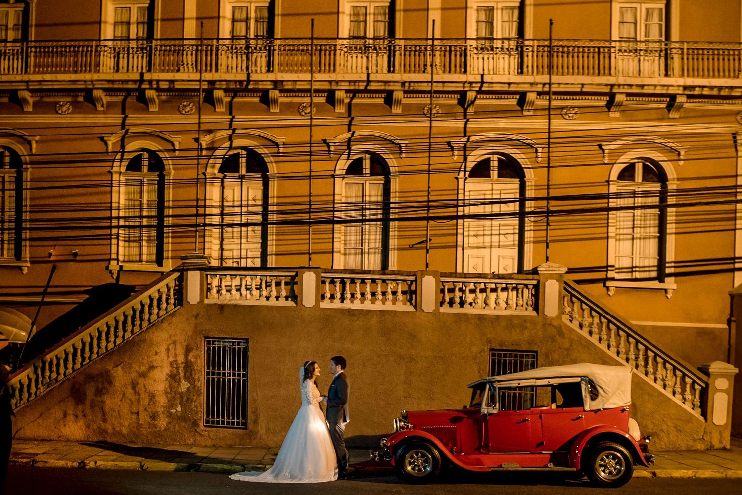 Vintage red car parked in front of illuminated yellow building with couple at night.