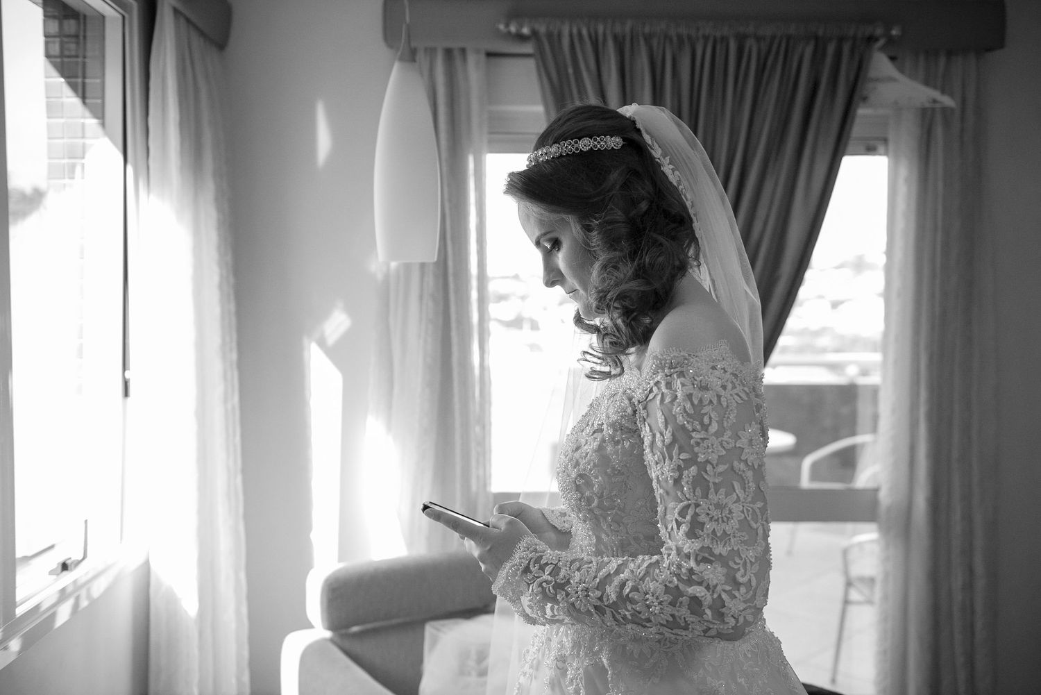 A bride in lace dress checks her phone by a sunlit window.
