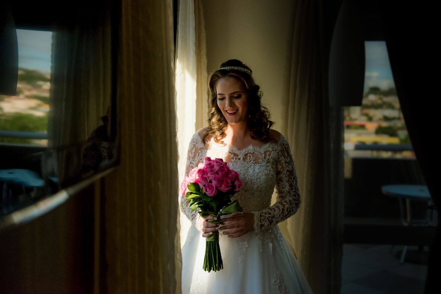 A bride in lace gown holds pink roses while standing in dramatic window light.