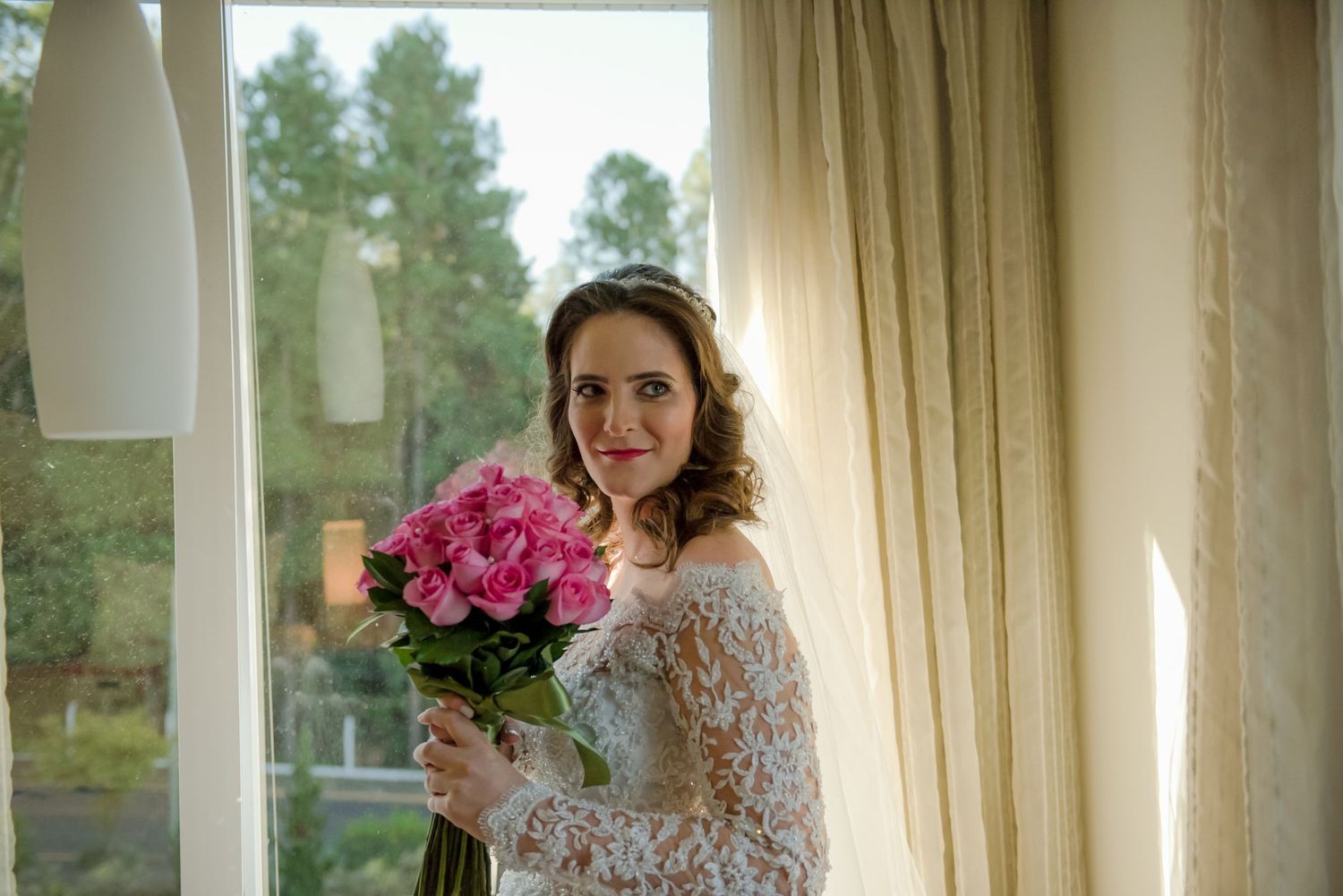 A bride in lace holding pink flowers poses by a window.