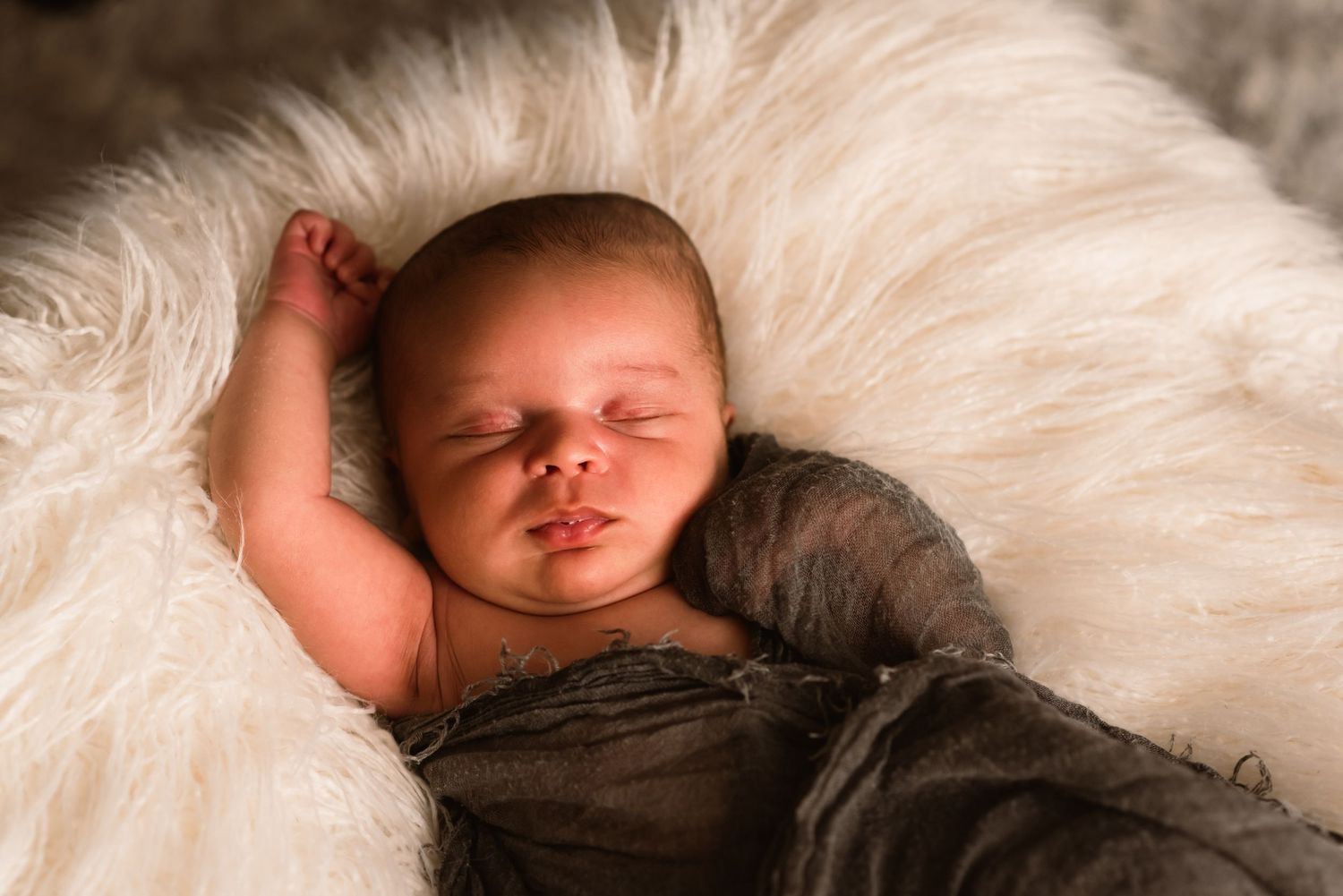 An image of a newborn baby with his arm sprawled out and gently wrapped in a grey textured blanket in this indoor newborn photoshoot