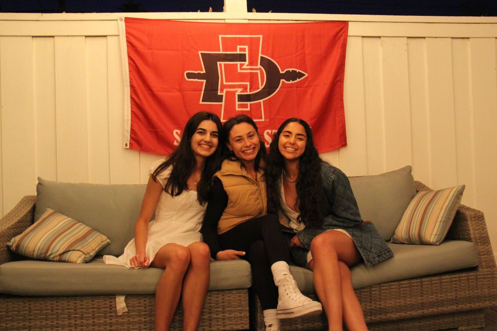 Students celebrate on outdoor patio furniture beneath red university flag.
