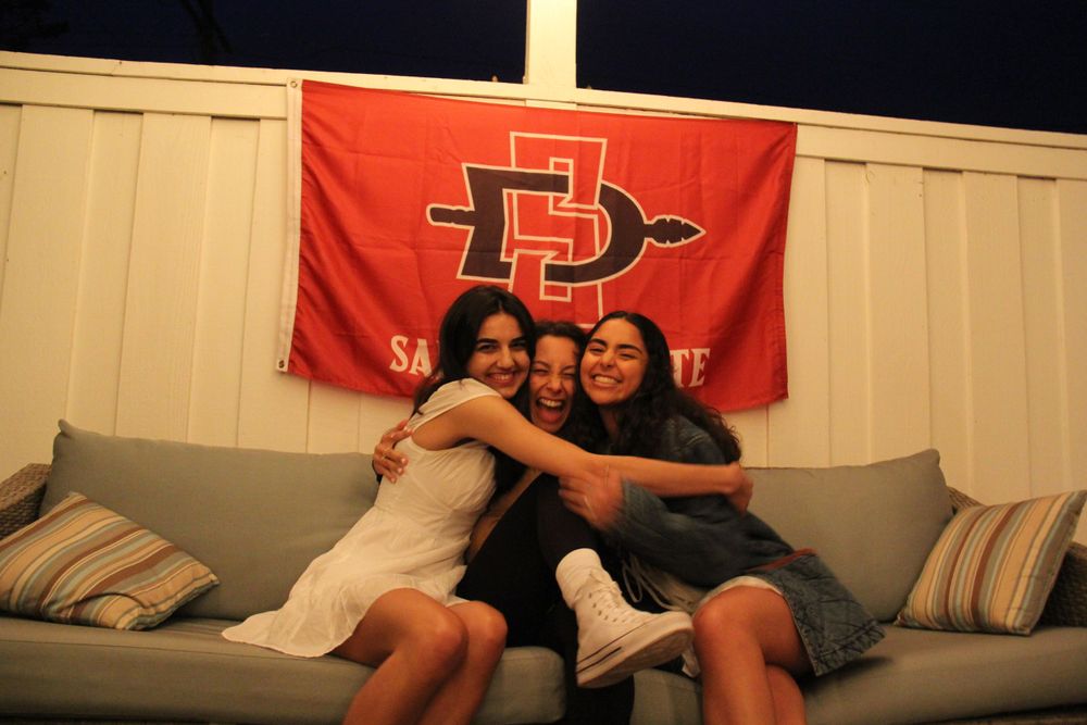 Friends embrace on gray couch beneath red university banner during celebration.