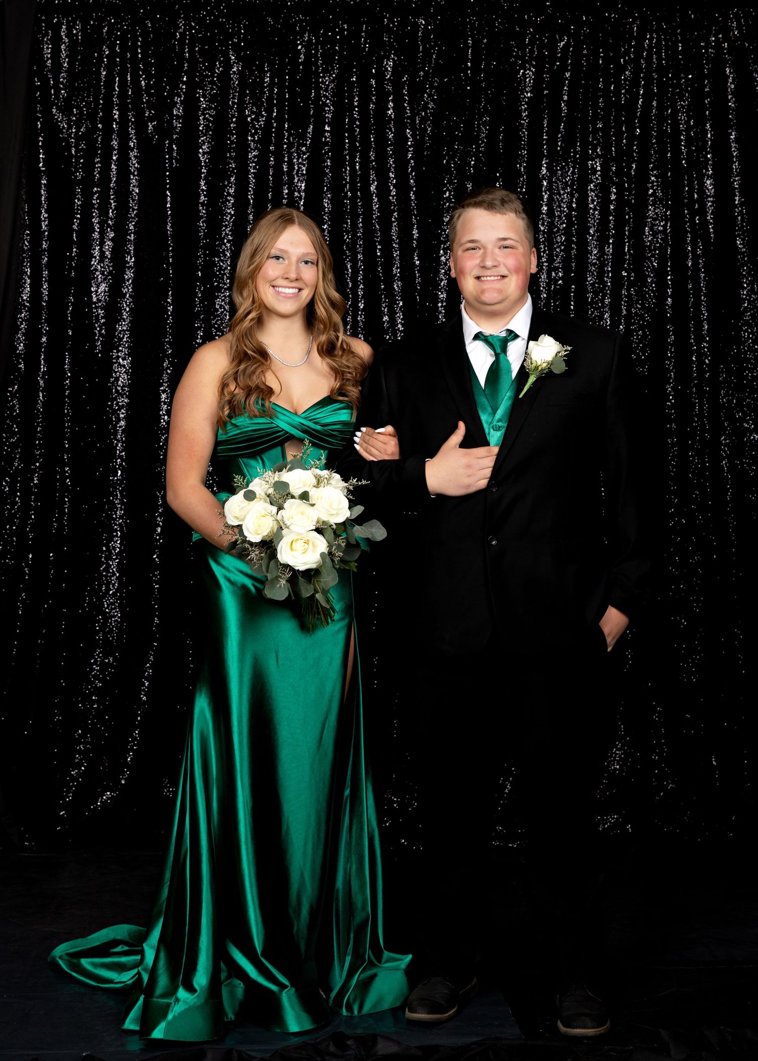 Couple in formal attire pose together, woman in emerald green gown holding white roses bouquet.