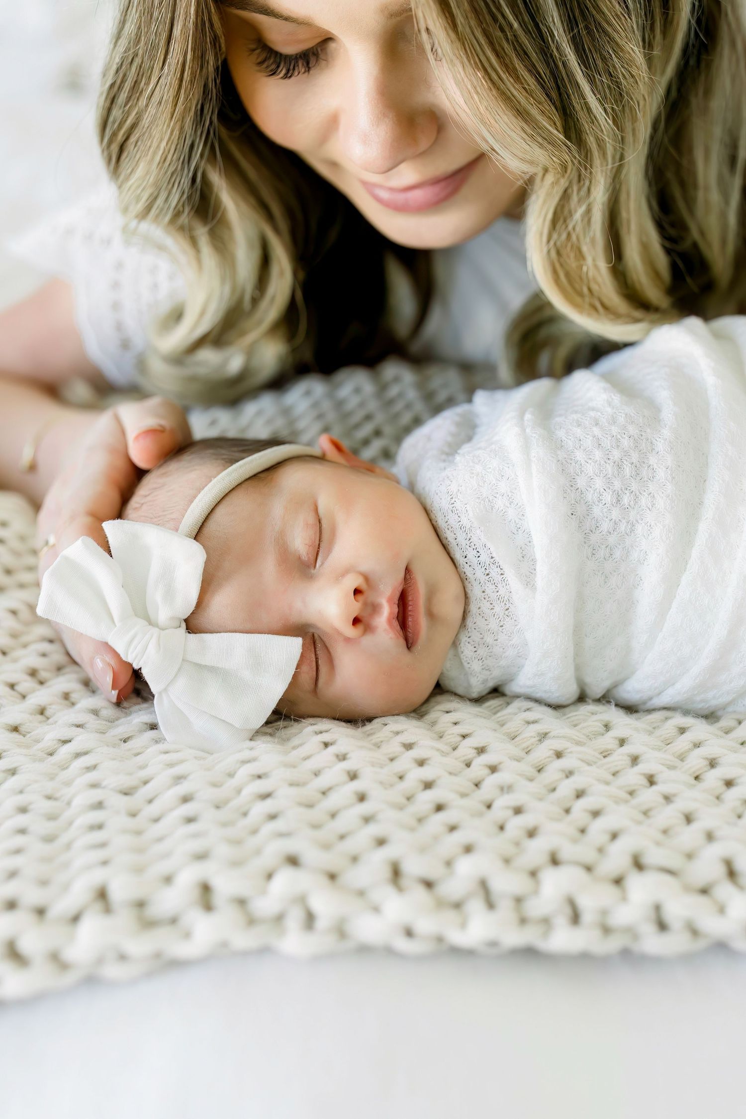 Sleeping newborn baby with white bow headband on knitted blanket.