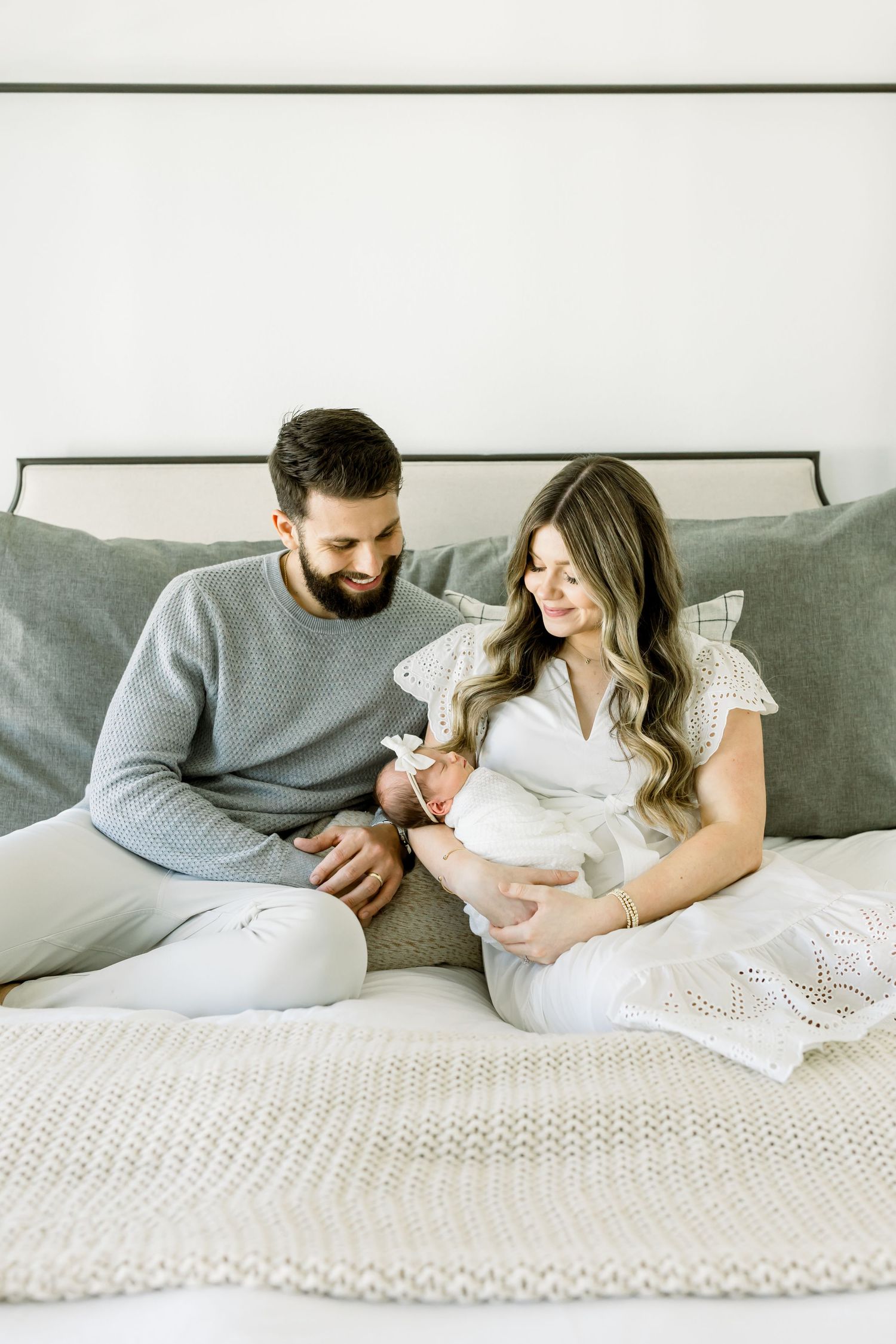 Parents sitting on bed with newborn baby, wearing matching grey and white outfits.